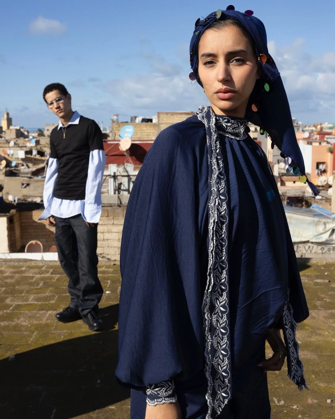 A woman with a headscarf decorated with colorful sequins standing in front, with a man in the background on a rooftop with city buildings and a blue sky.