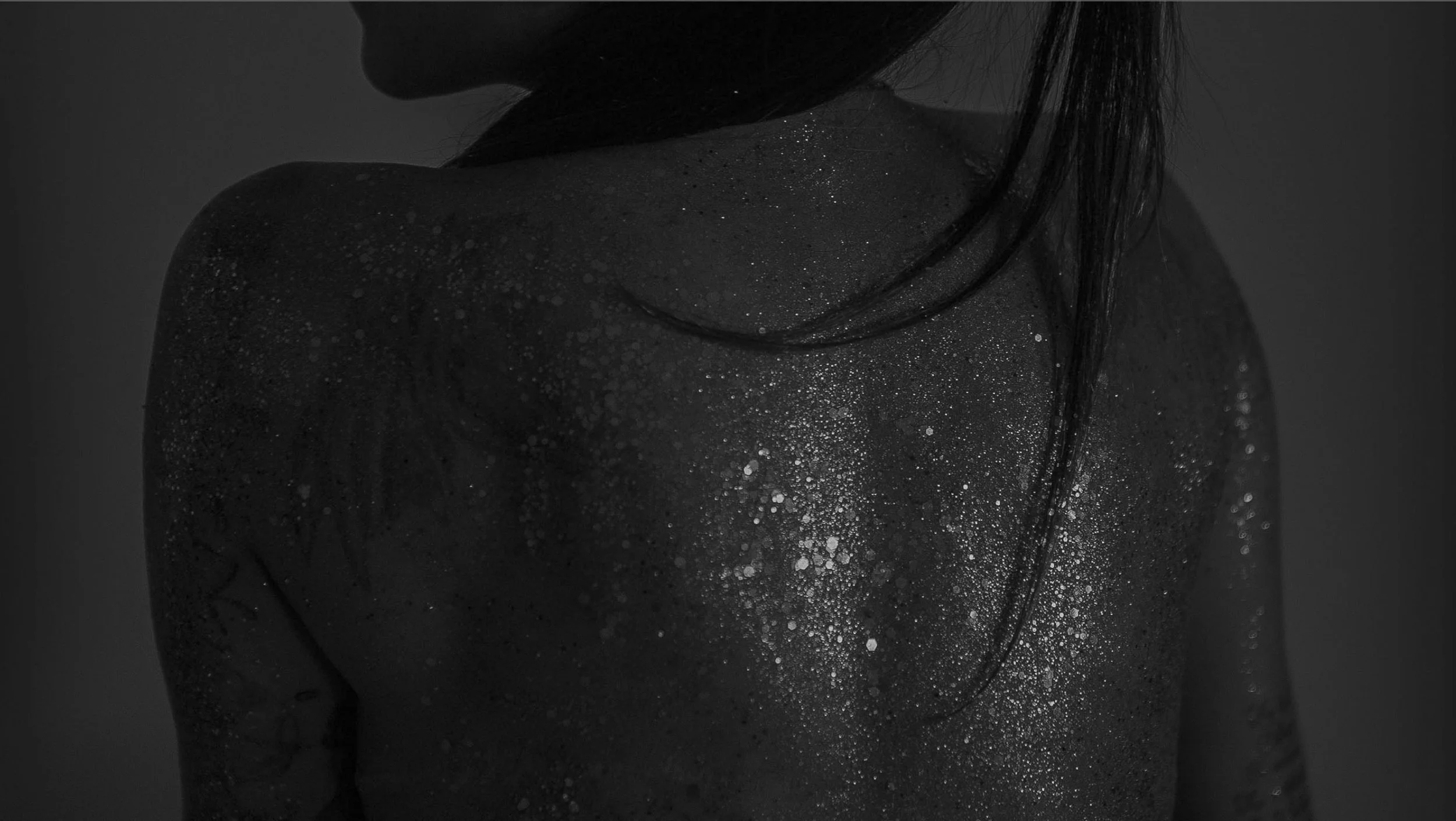 Close-up of a person's shoulder, neck, and chest, covered in glitter on dark skin, with long dark hair partially covering the face, in black and white.
