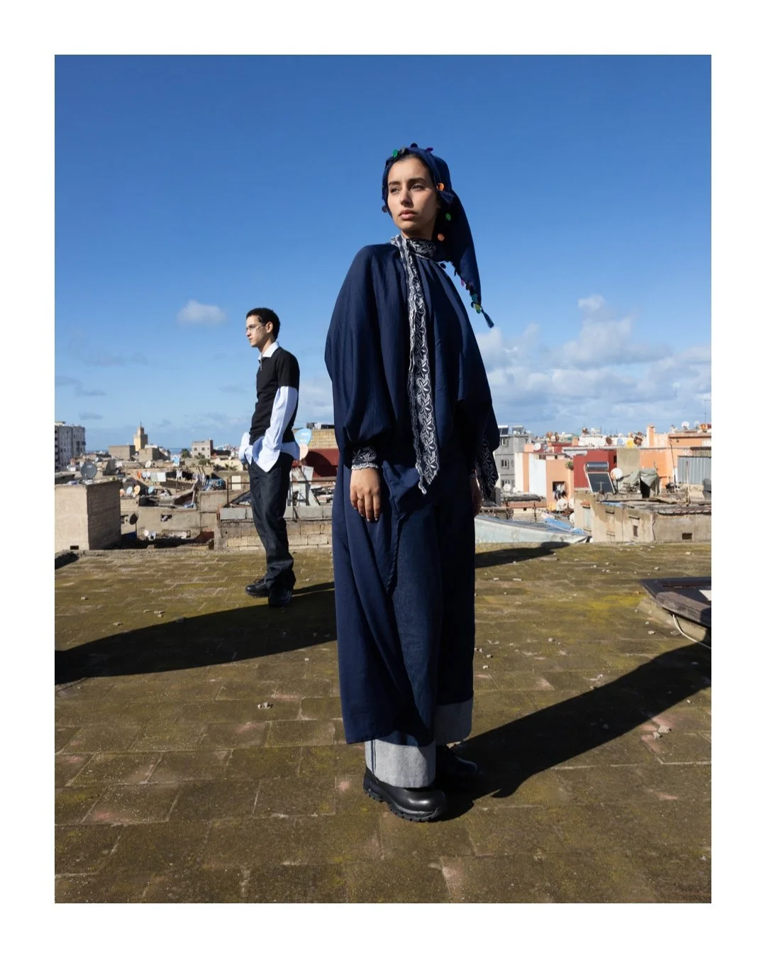 Two individuals stand on a rooftop with a cityscape in the background under a clear blue sky. The woman in the foreground wears a blue headscarf, loose blue clothing, and black boots. The man in the background wears a black sweater with white shirt s