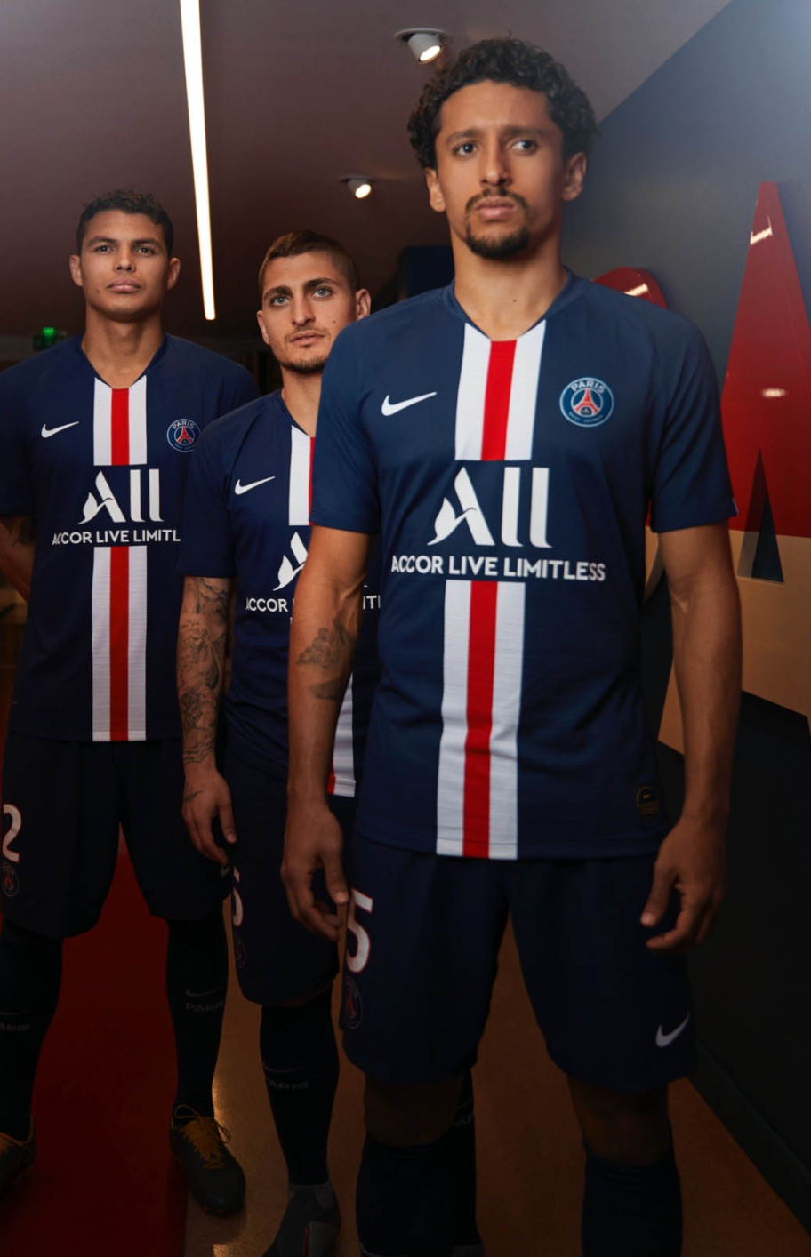 Four male soccer players standing indoors, wearing matching Paris Saint-Germain soccer uniforms with blue jerseys, red and white vertical stripes, and the club's logo. They are looking serious, with one slightly in front of the others.