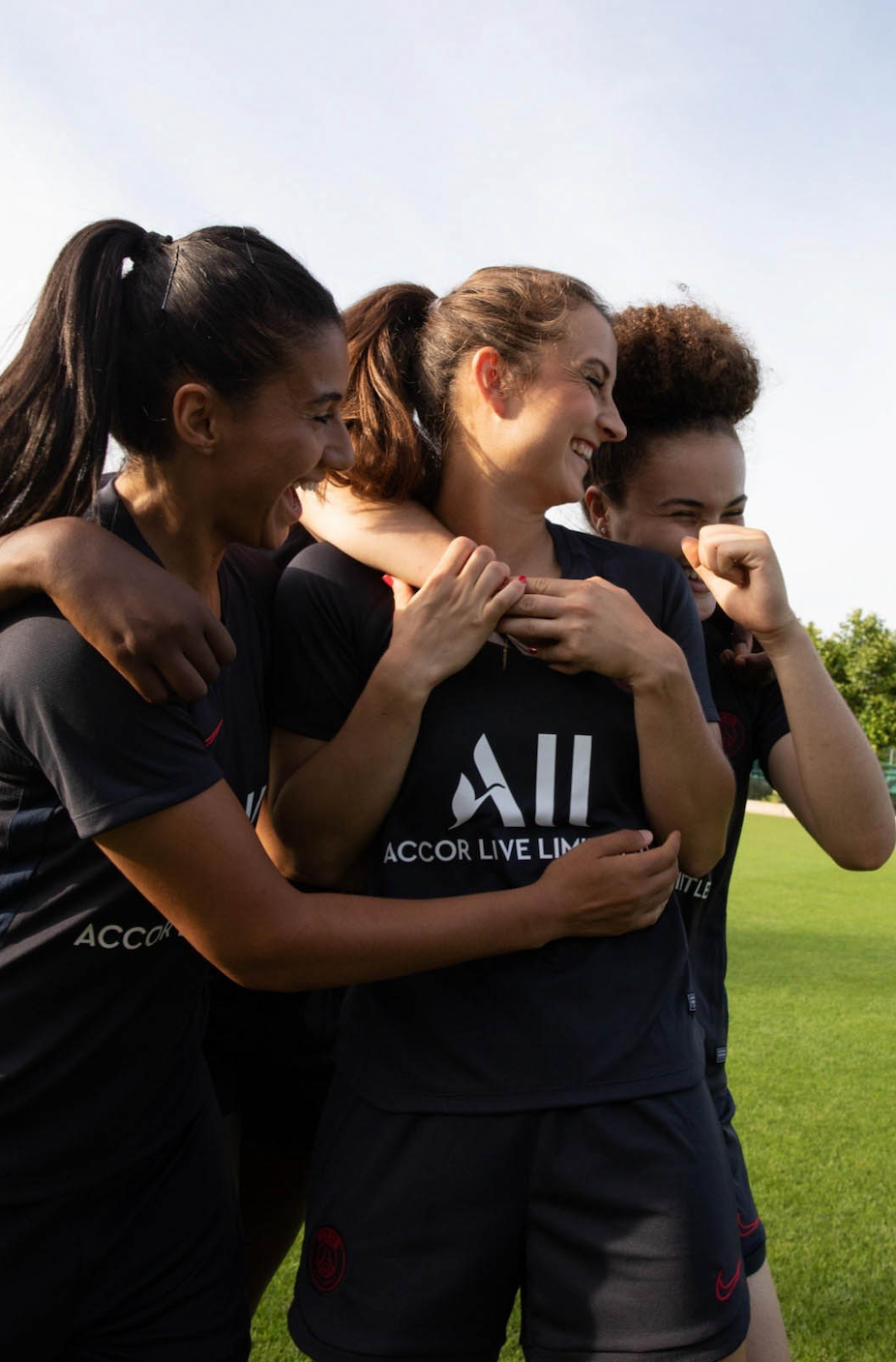Group of four female soccer players celebrating on the field, laughing and hugging each other, wearing black jerseys.