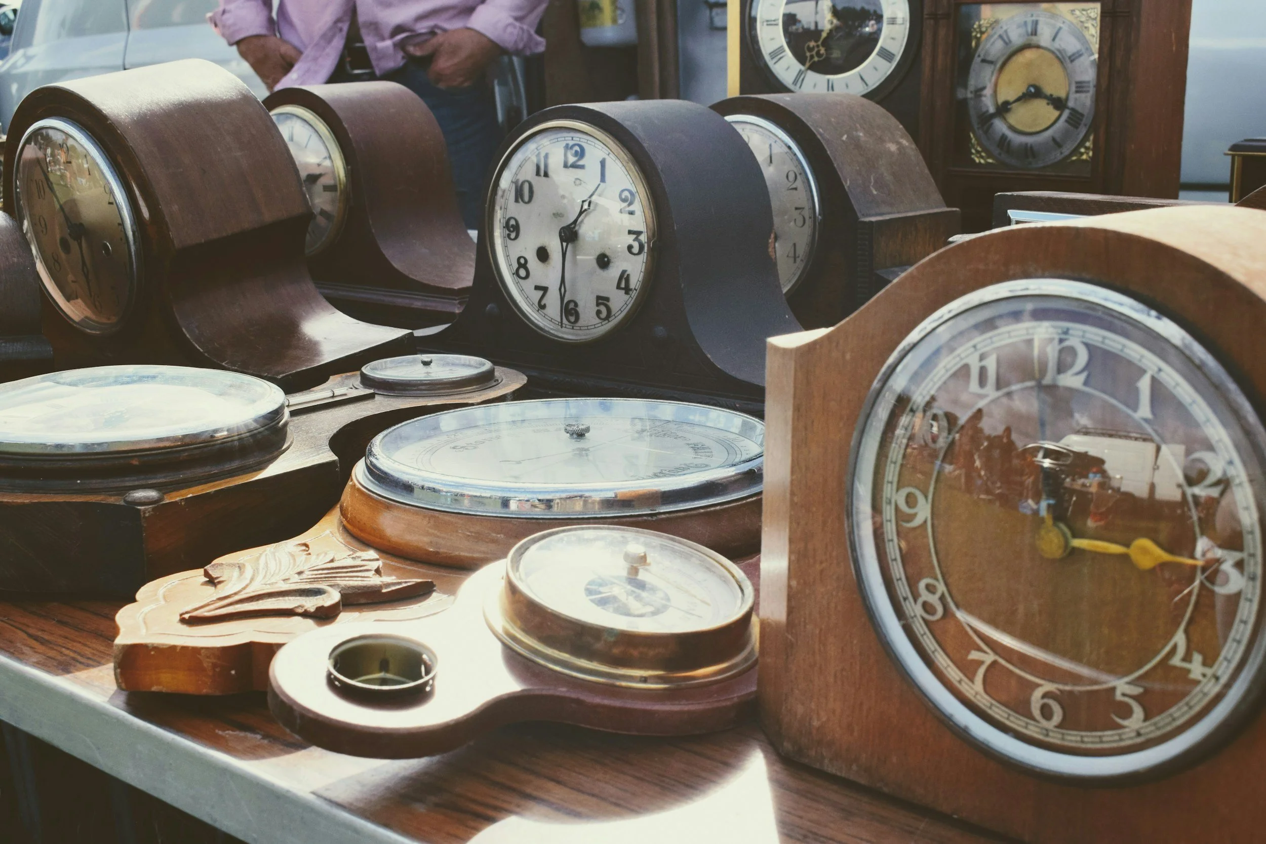Multiple vintage clocks and clock parts displayed on a table at an outdoor market.