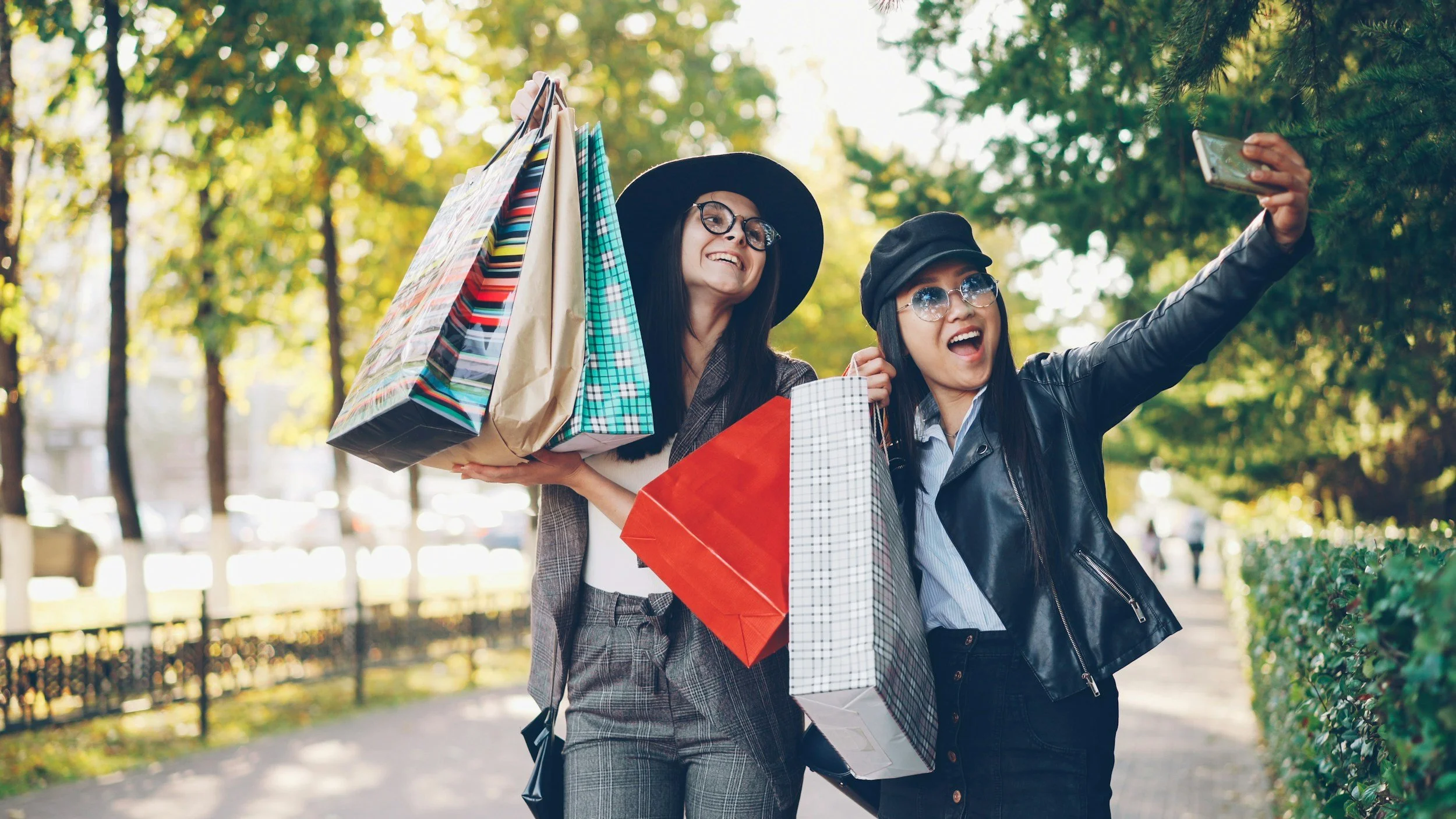 Two women with shopping bags taking a selfie outdoors in a park during fall