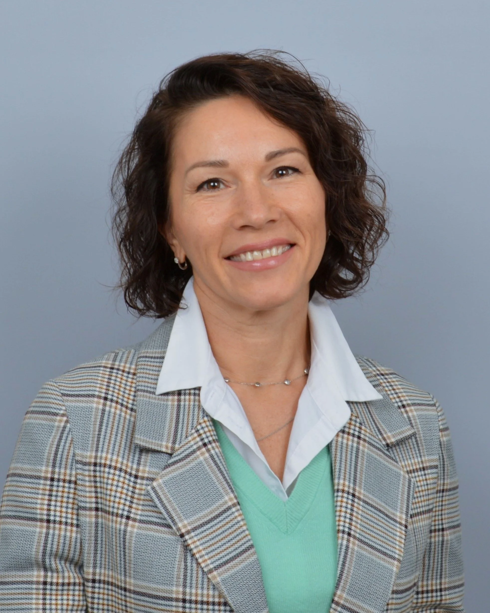 A woman with curly dark brown hair smiling against a plain gray background. She is wearing a chequered blazer, a white shirt, a light green sweater, and small earrings.