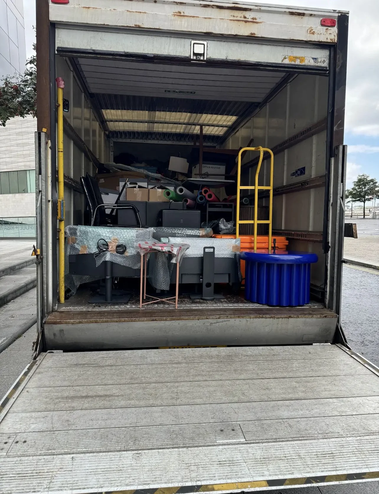 The back of a moving truck filled with office furniture and supplies, including chairs, boxes, and plastic storage bins.
