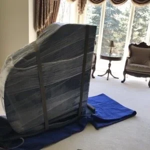 Disassembled bed frame wrapped in plastic and placed on a blue cloth in a living room with large window and curtains.