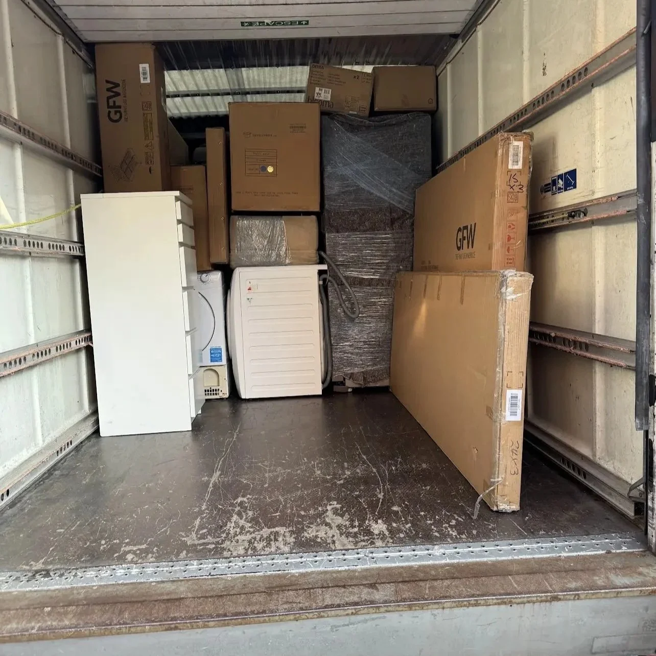 Inside a truck, with various boxes and appliances stored, including a white mini-refrigerator, washing machine, and large flat boxes in cardboard packaging.