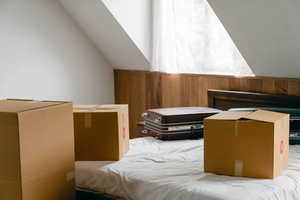A bed with four cardboard boxes and three hard-shell suitcases placed on it, in a room with a sloped ceiling and a window with sheer curtains.