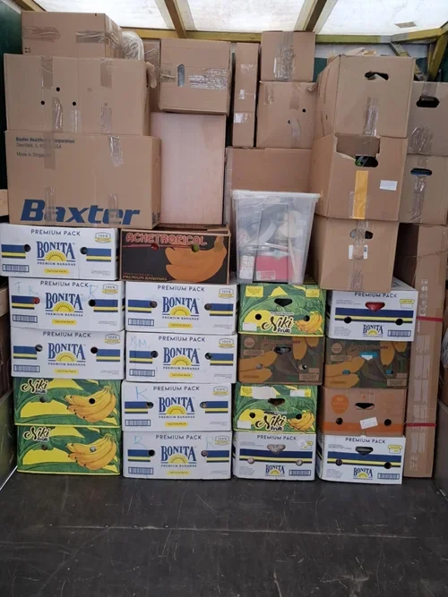 Stacked boxes of various brands and types of fruit, including boxes of bananas and other produce, inside a storage area or warehouse.