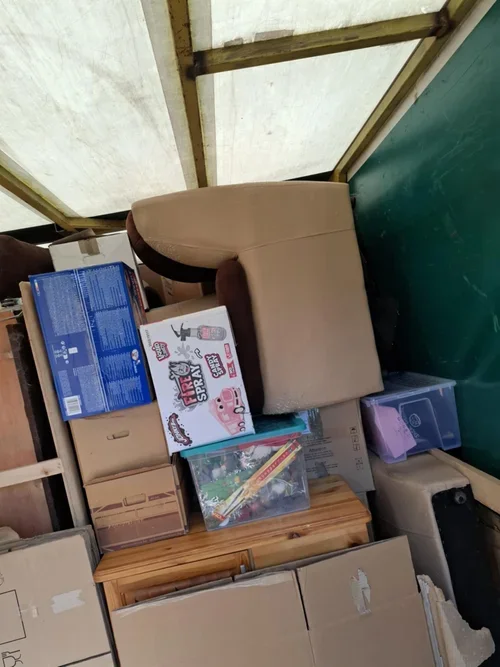 A beige armchair surrounded by various boxes and items, with a wooden crate and a clear plastic container on the floor.