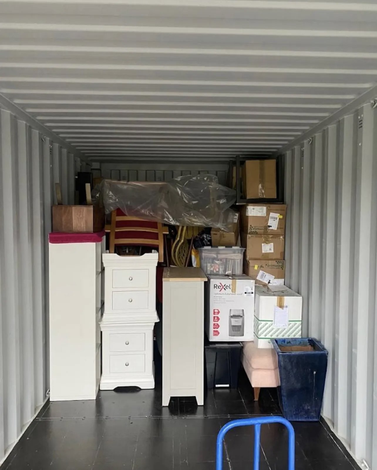 The image shows the inside of a storage unit filled with various furniture and boxes, including a white chest of drawers, a pink ottoman, a black storage bin, and several cardboard boxes stacked on top of each other.