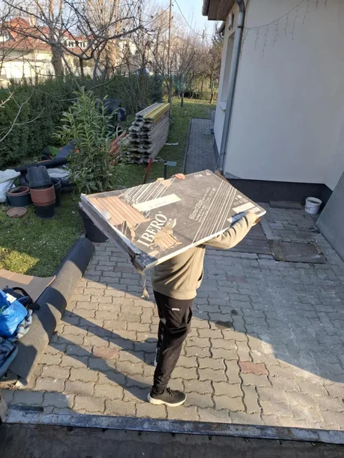 Child carrying a large cardboard box outside on a paved area next to a house with garden supplies and plants in the background.