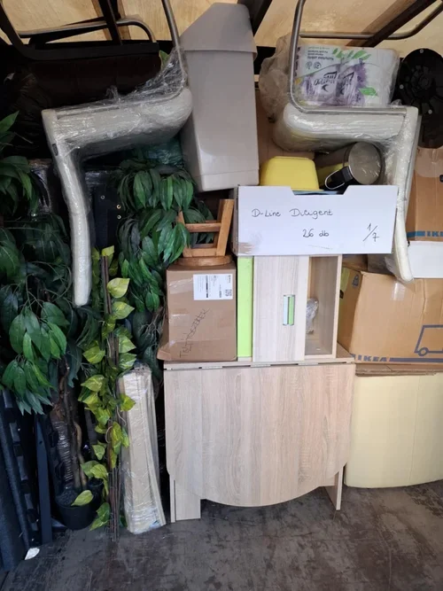 A cluttered storage area with assorted furniture pieces, plants, boxes, and miscellaneous items piled on and around a small wooden cabinet.