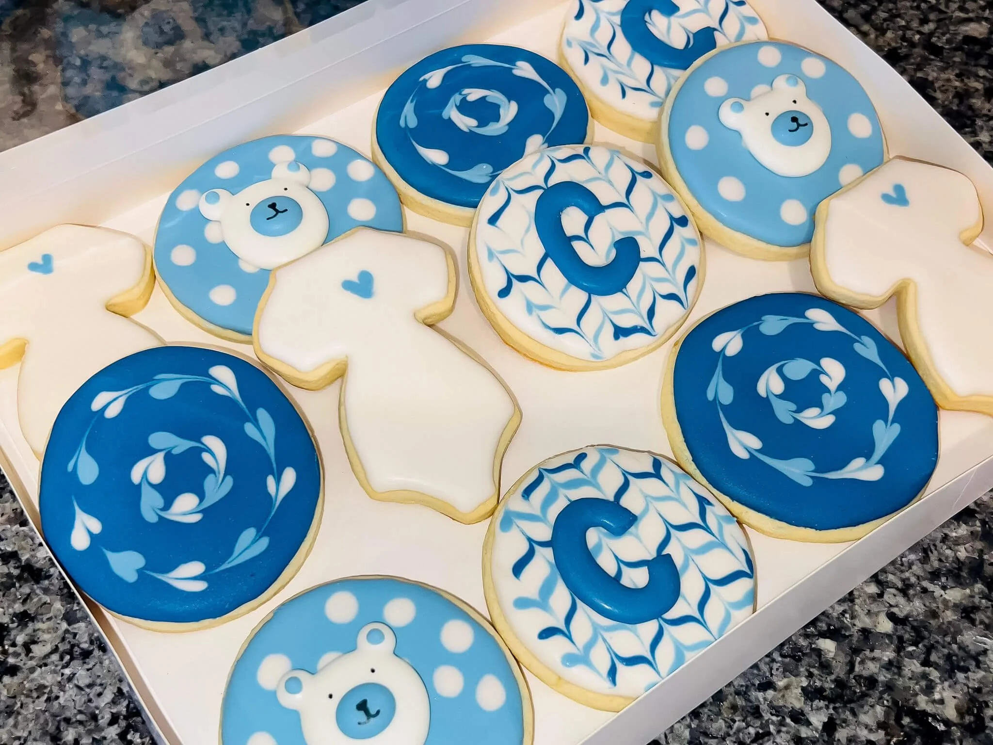 A collection of decorated cookies with a blue and white color theme, featuring bear faces, the letter 'C', and various swirls and patterns, arranged on a white tray.