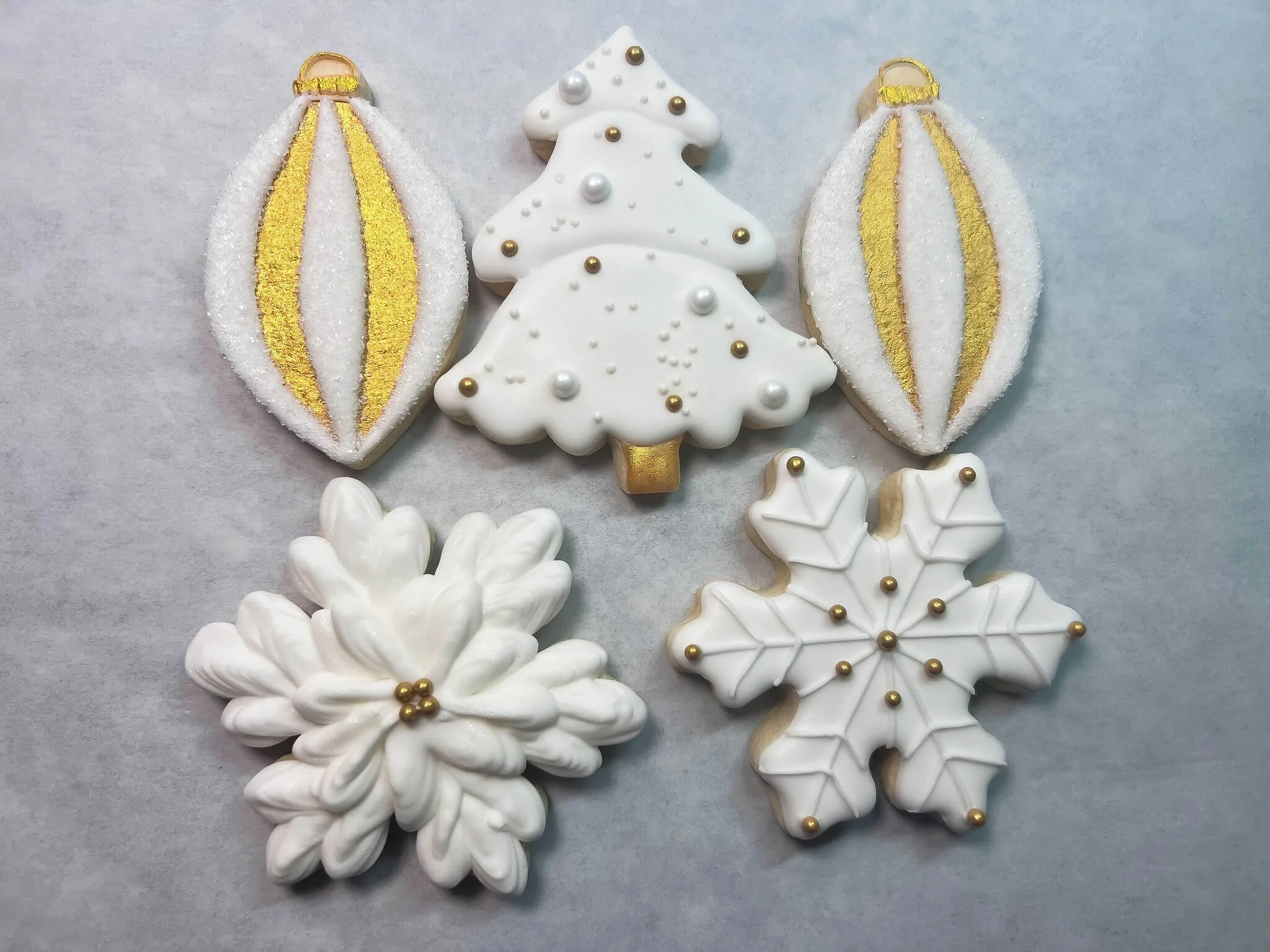 Festive holiday cookies including two gold and white oval ornaments, a white Christmas tree, a white snowflake, and a white flower, all decorated with white and gold icing and beads.