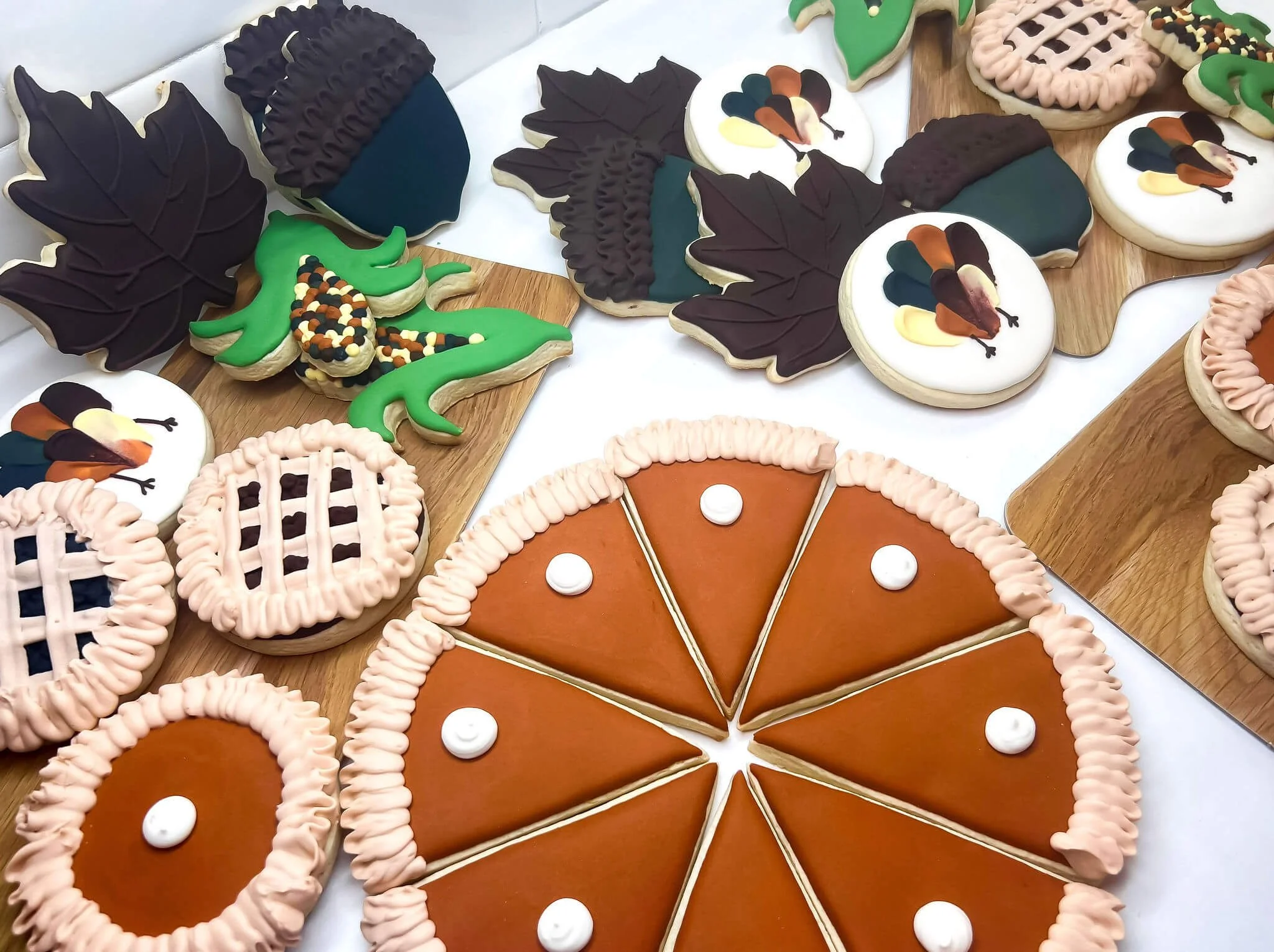 Assorted decorated sugar cookies including a pumpkin pie, a cornucopia, autumn leaves, a turkey, a gourd, a cornucopia filled with colored candy, and a turkey with its feathers spread, all on wooden and white surfaces.