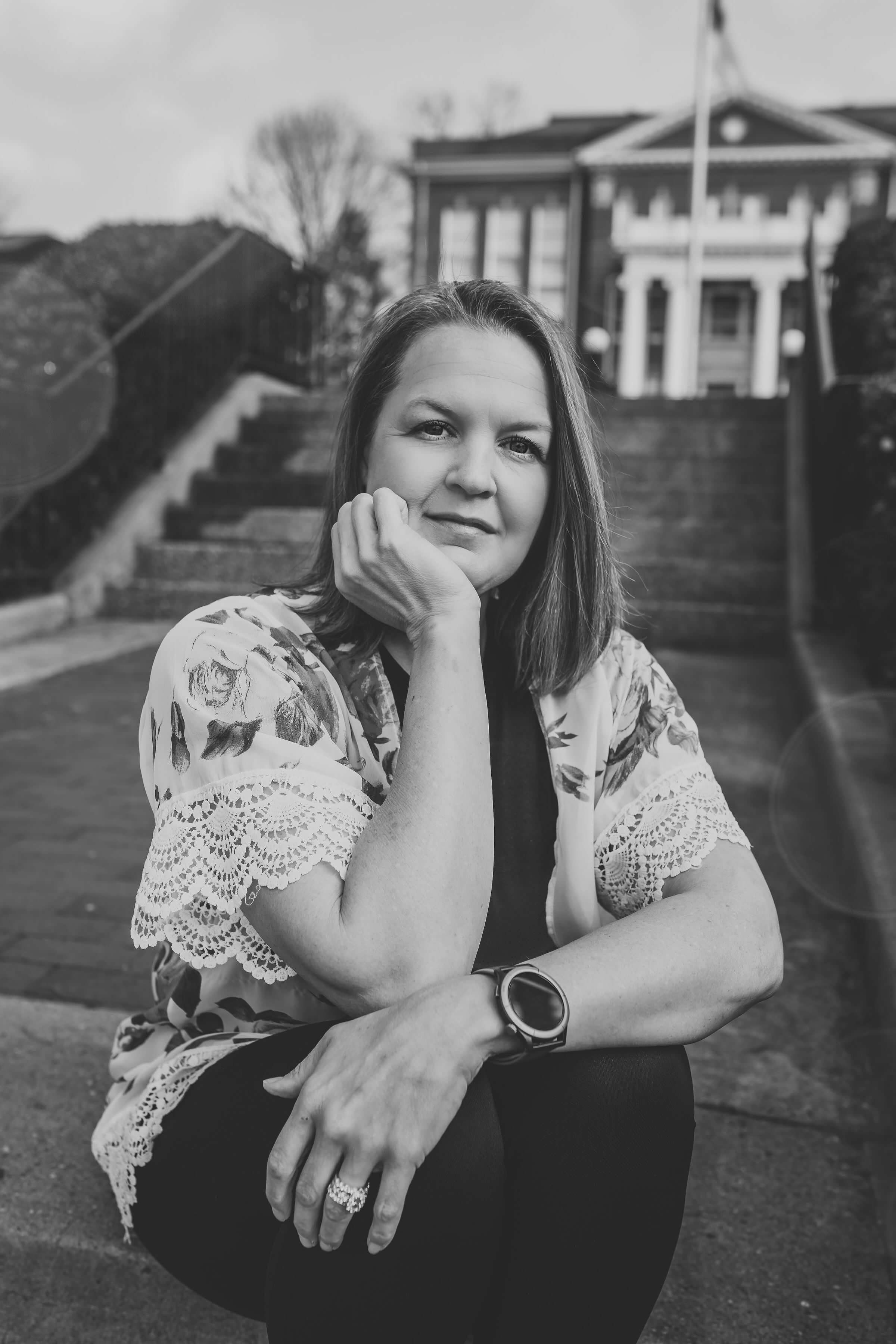 A woman with shoulder-length hair sitting outdoors with a hand resting on her chin, wearing a floral-patterned blouse with lace details and a smartwatch, in front of a building with stairs and a flagpole.