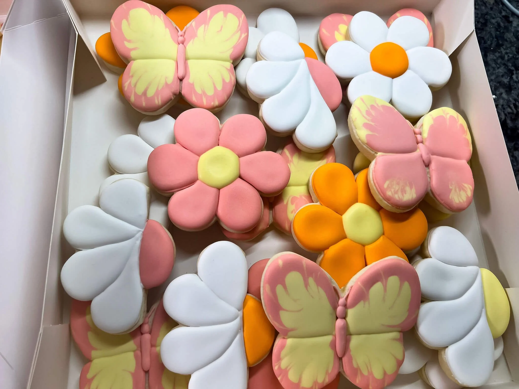 Assorted decorated cookies in butterfly, flower, and petal shapes with pink, white, yellow, and orange icing, arranged in a white box.