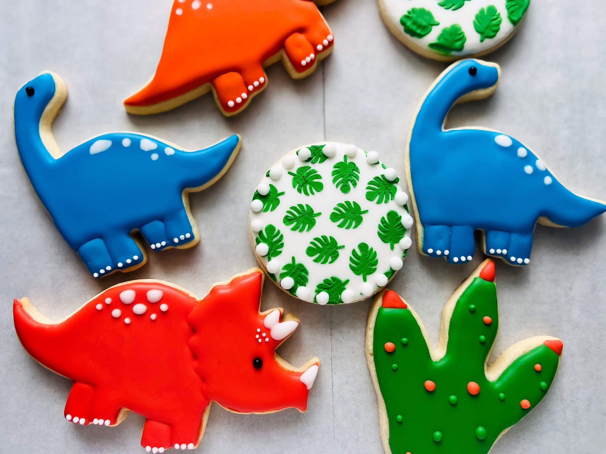 Colorful dinosaur-shaped cookies decorated with bright icing in blue, red, and orange, and round cookies with green leaf patterns and sprinkles.
