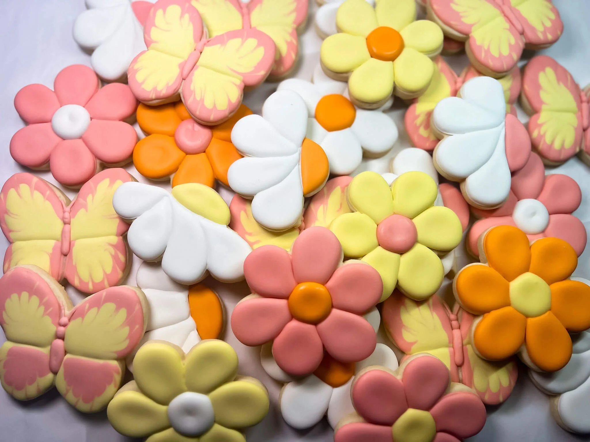 Colorful flower and butterfly-shaped sugar cookies decorated with pastel icing in pink, yellow, white, and orange.