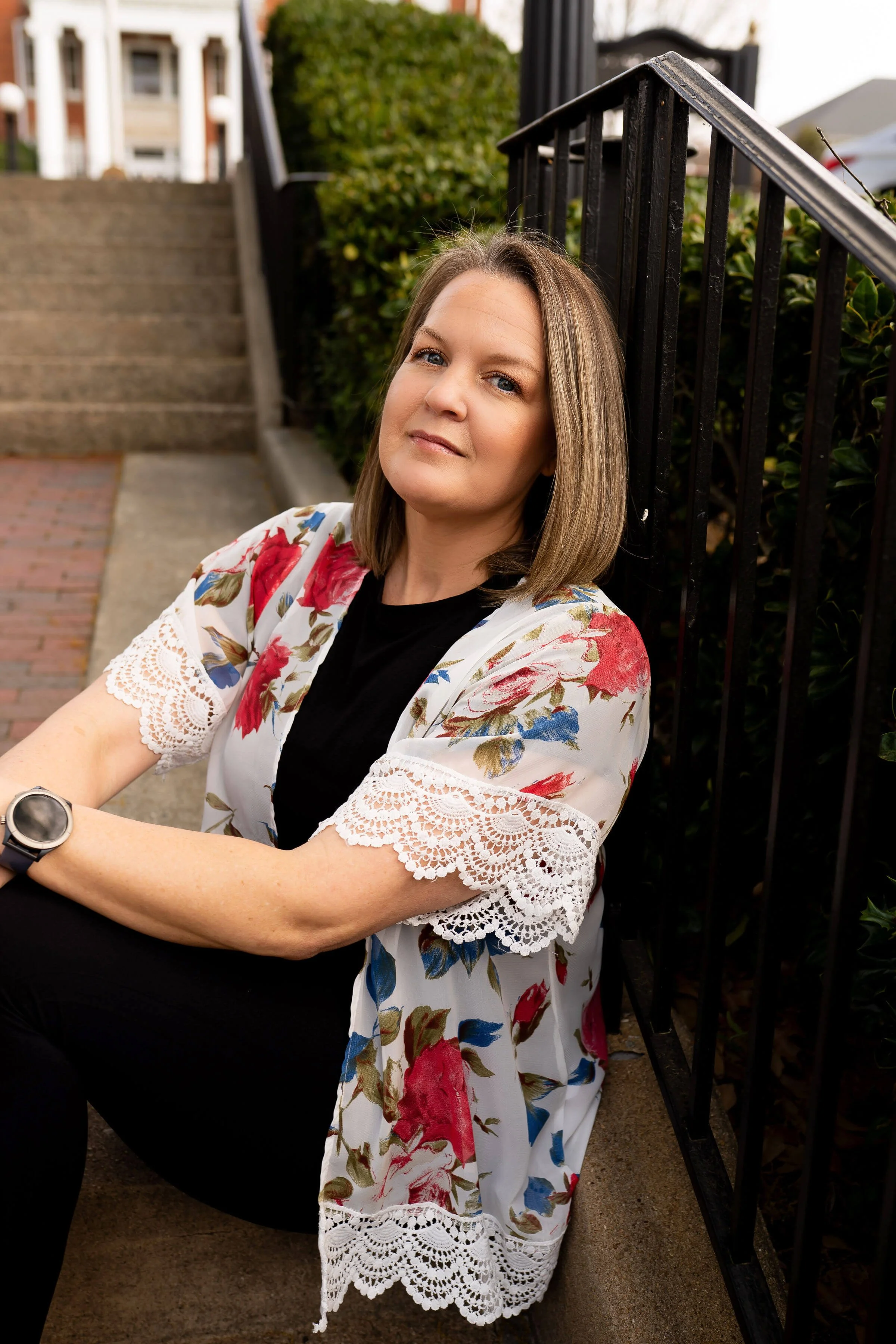 A woman with shoulder-length light brown hair sitting on a step near a black metal fence, wearing a white floral kimono with lace trim over a black top, with a smartwatch on her left wrist, and looking at the camera.