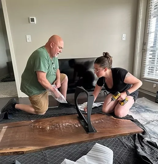 Two people, a man and a woman, installing a large wooden tabletop on a black metal base in a living room.