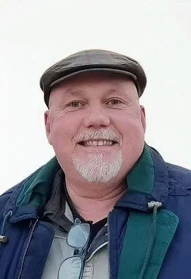 Smiling man with a gray beard and mustache wearing a hat, glasses hanging from his shirt, and a blue jacket against a white background.
