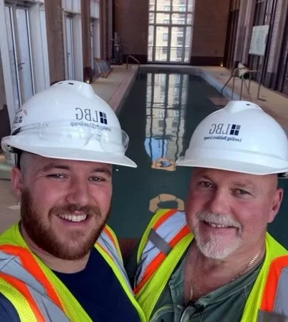 Two men wearing hard hats and safety vests smiling inside a swimming pool area.
