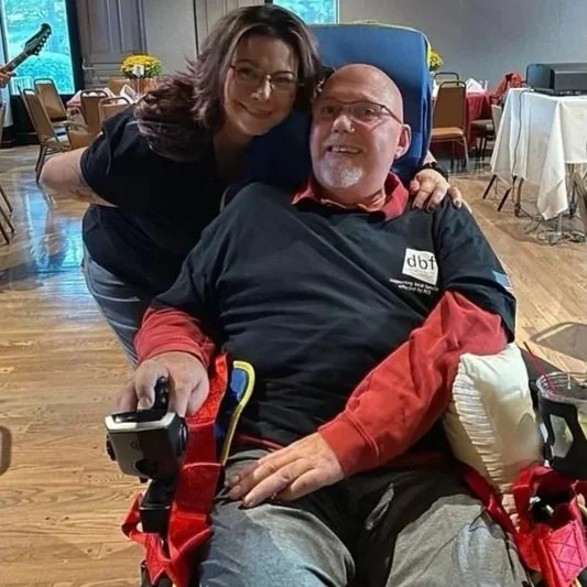 A woman standing next to a man in a chair with medical equipment, smiling at the camera in a decorated room with tables and chairs.