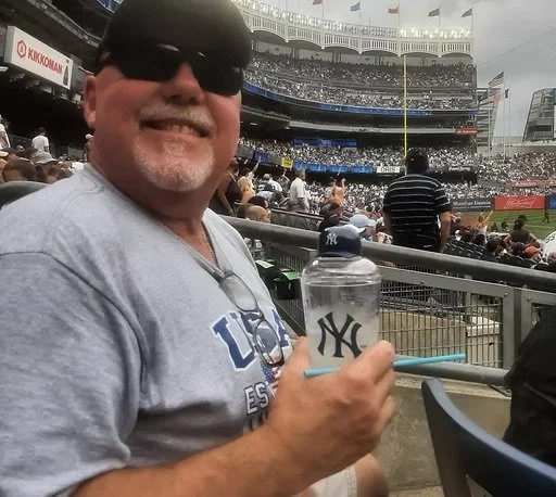 Man at baseball game sitting in stands, smiling, wearing sunglasses, holding a transparent water bottle with the New York Yankees logo, and a light blue pen, with a stadium filled with spectators in the background.