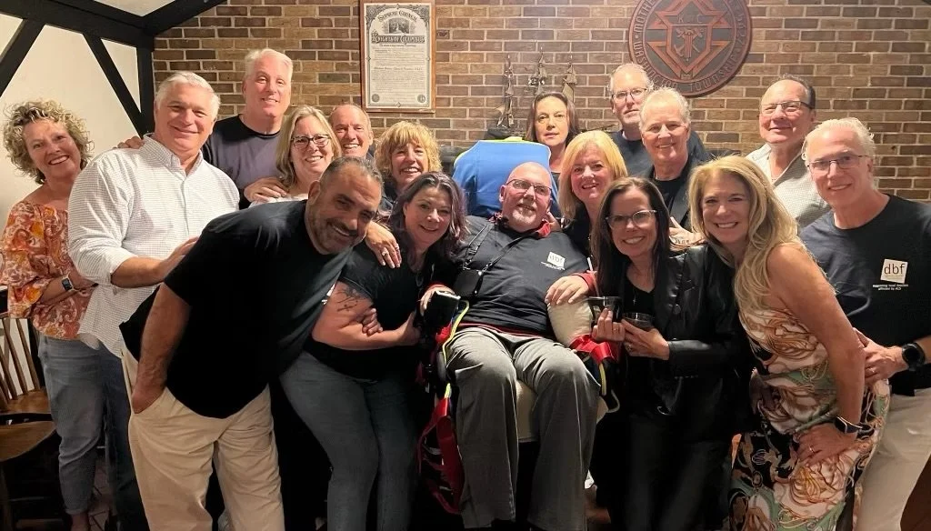 Group of 14 people, including a man in a wheelchair, celebrating together in a brick room, smiling and posing for the photo.
