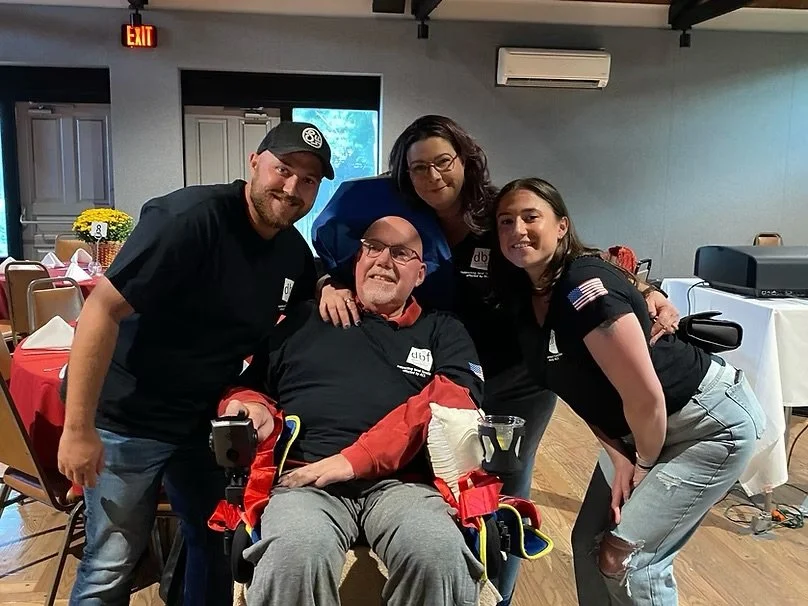 Four people smiling around a man in a wheelchair, with two women and one man standing close, all wearing black shirts. The setting is an indoor room with decorated tables and chairs, suggesting a social or celebratory event.
