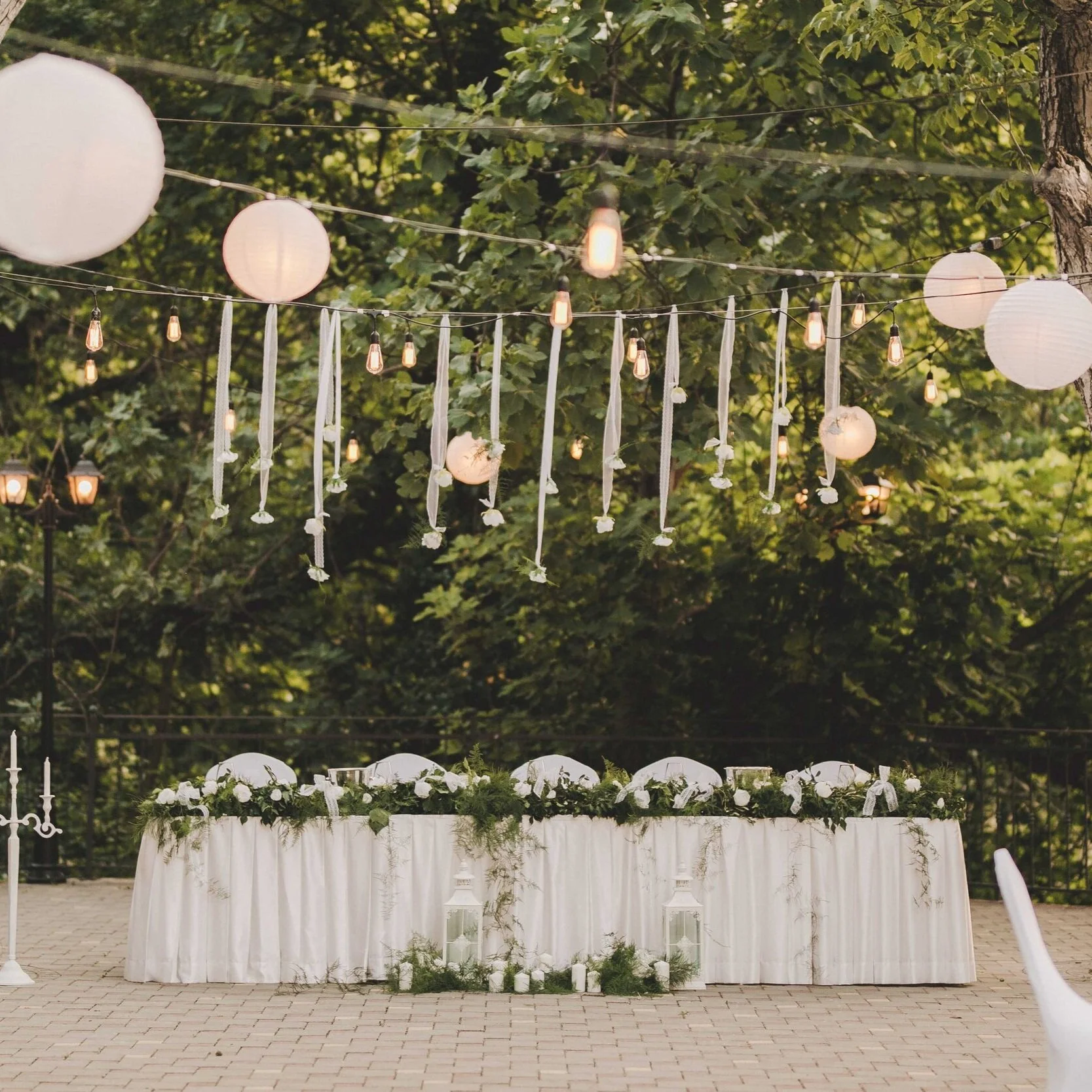 Ein festlich gedeckter Tisch mit weißen Tischtüchern, Blumen und Kerzen, im Hintergrund ein Barbecue-Bereich, darüber hängen Papierlaternen und Lichterketten, alles im Freien bei Baum- und Grünflächen.