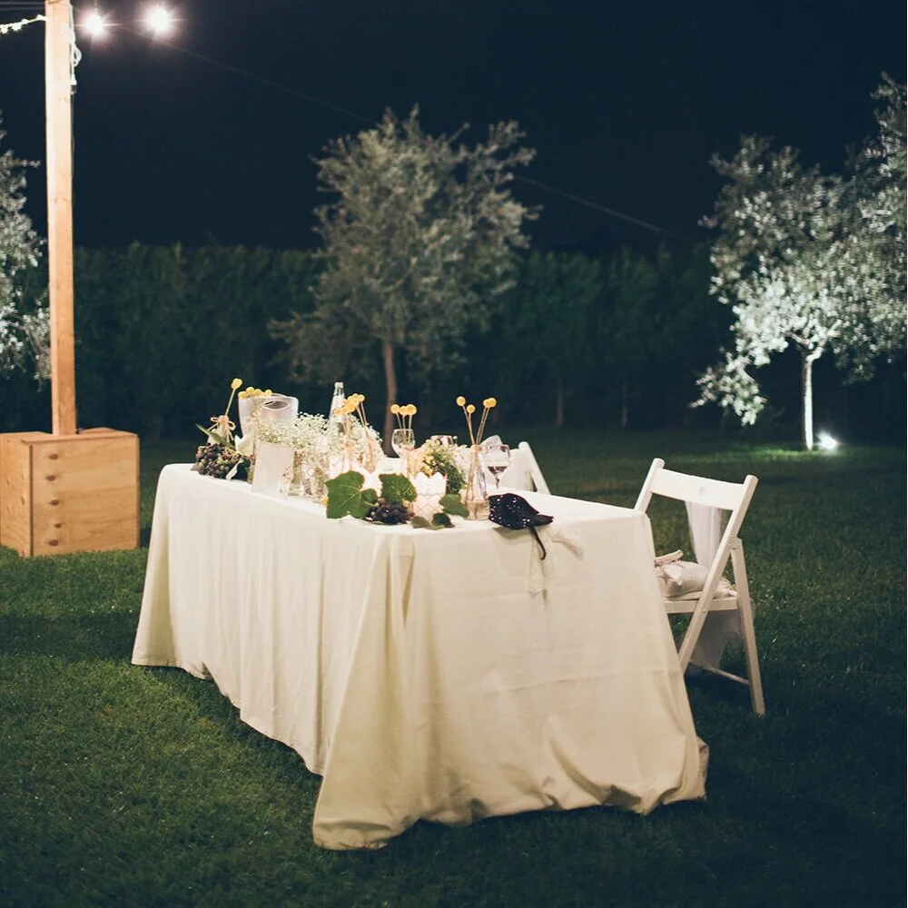 Ein im Freien gedeckter Esstisch bei Nacht mit Wein, Gläsern, Blumen und Dekorationen, umgeben von Bäumen und einem hell erleuchteten Himmel.