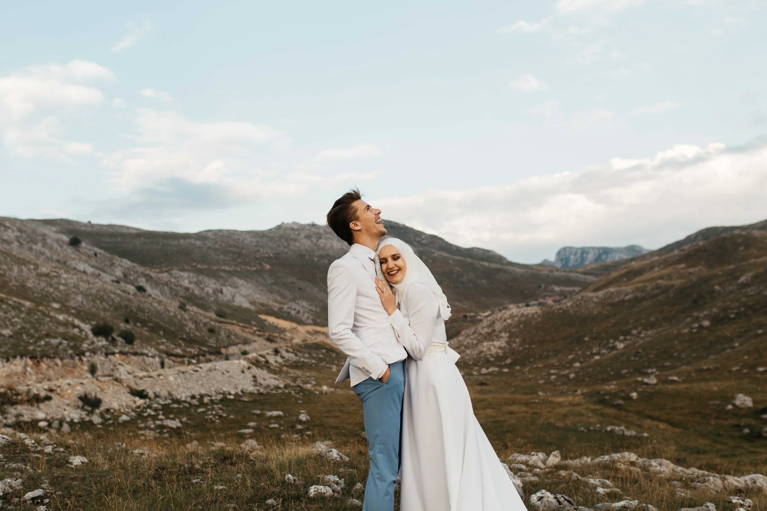 Ein lachendes Paar, wahrscheinlich bei einer Hochzeit, umarmt sich in einer bergigen Landschaft mit bewölktem Himmel.