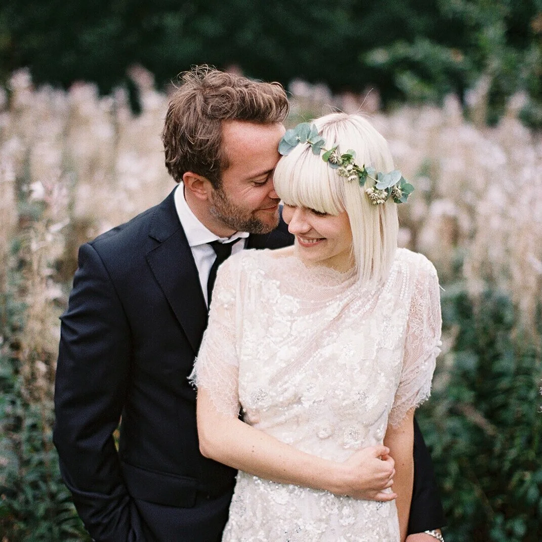 Ein verliebtes Paar bei einer Hochzeit, der Mann trägt einen schwarzen Anzug und die Frau ein weißes Spitzenkleid mit Blumenkronen im Haar, sie lachen im Grünen.