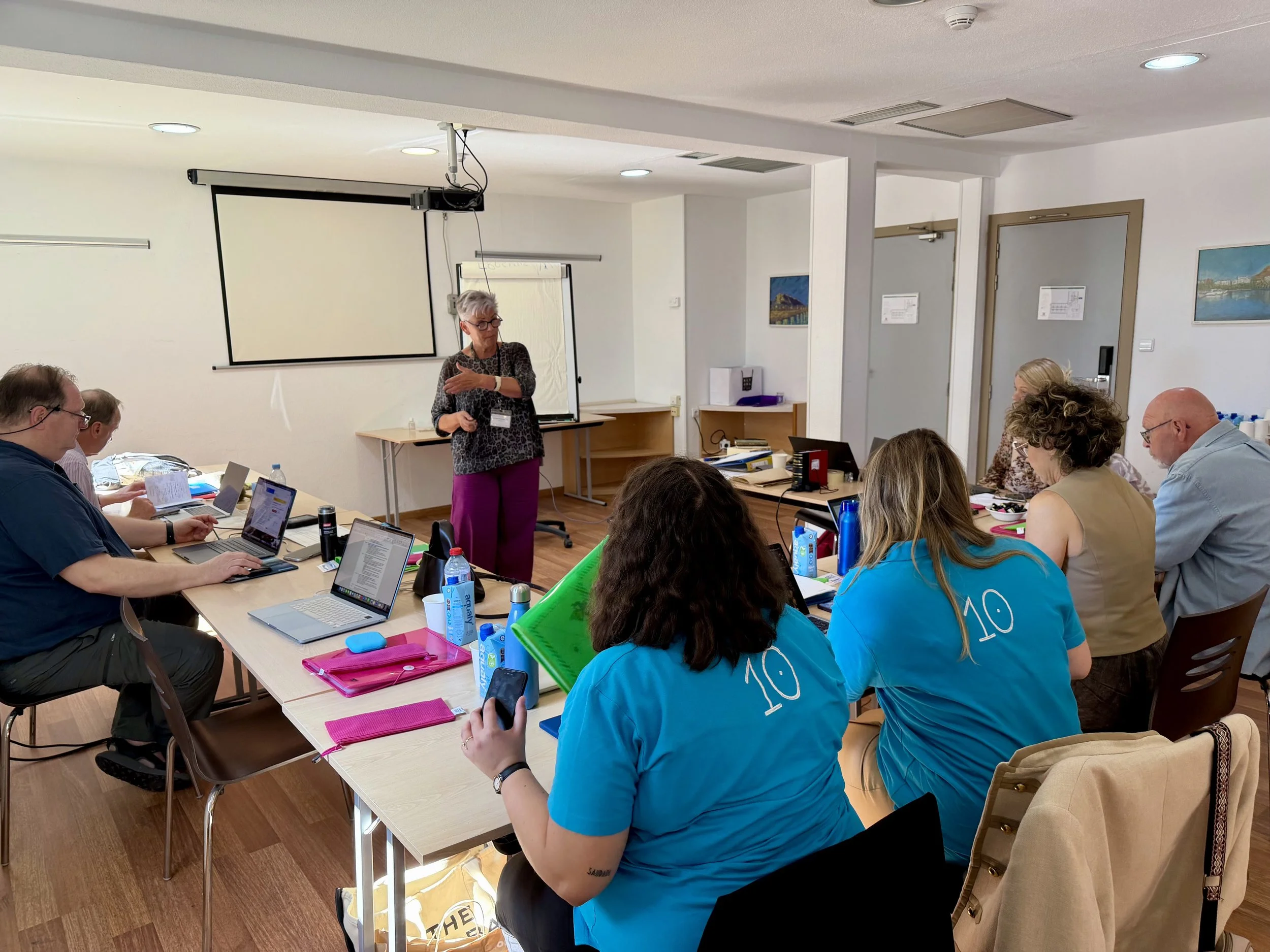 A woman standing and speaking to a group of people seated around a conference table in a meeting room with laptops and water bottles.