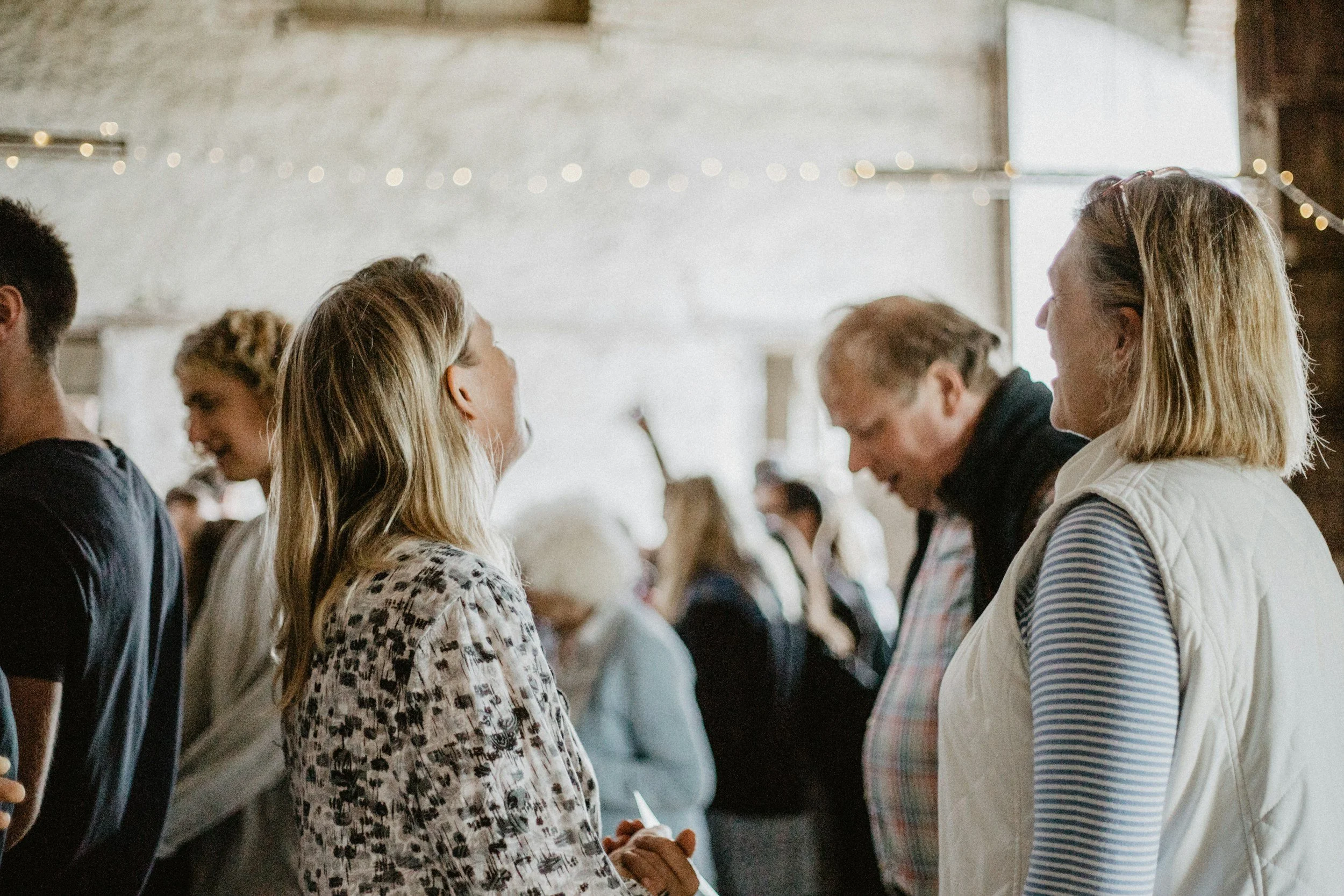 People talking and smiling in a social gathering inside a barn-like venue with string lights.