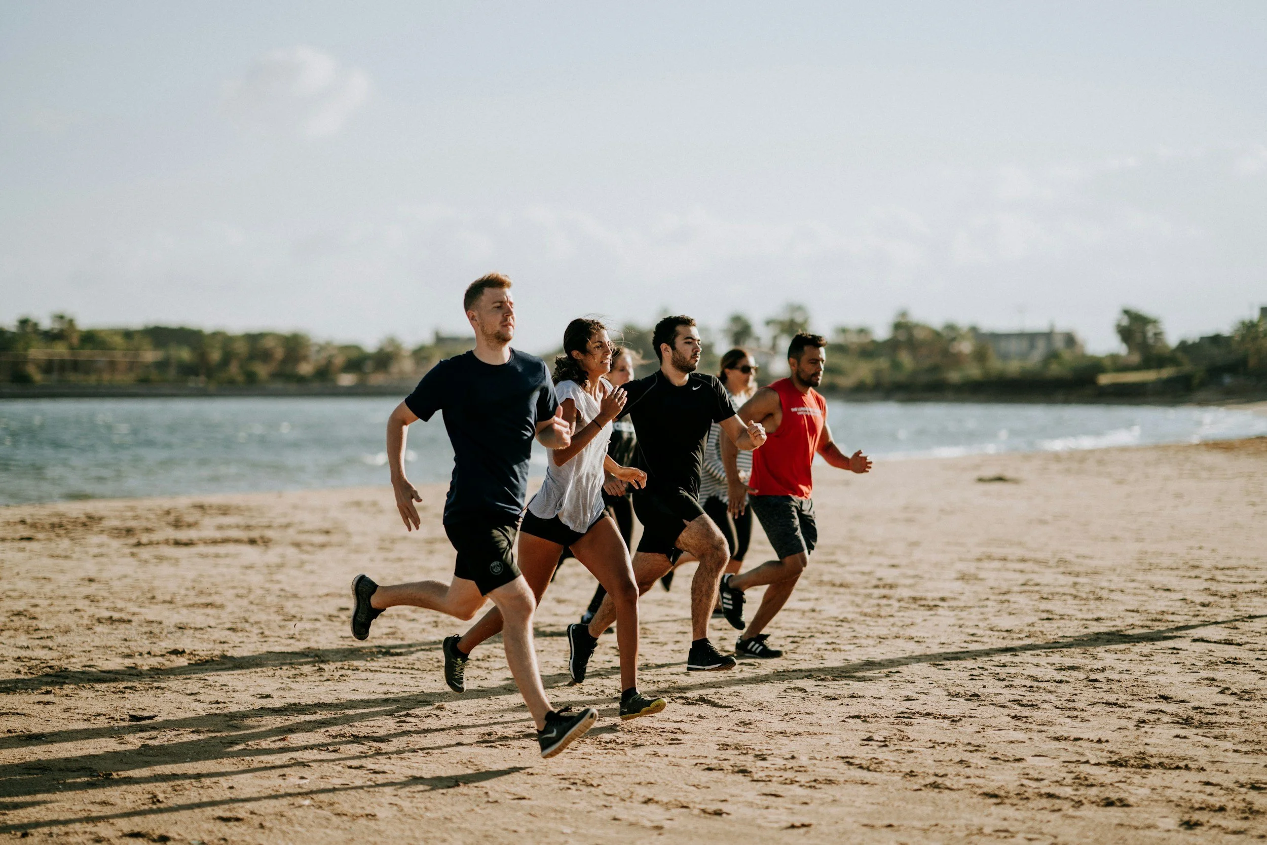 Group of six people running on a sandy beach near water during daytime.