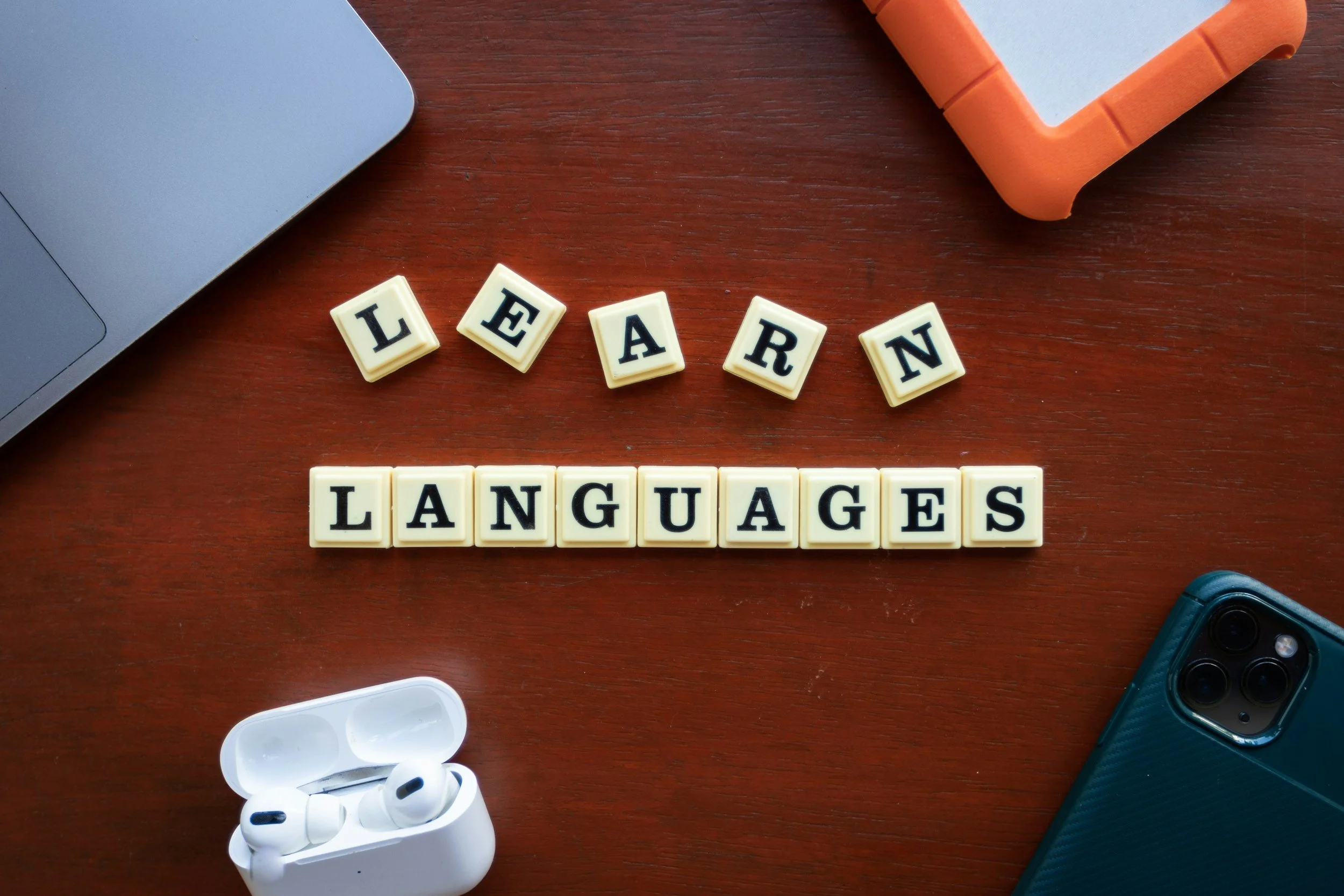 Scrabble tiles spelling out 'LEARN' and 'LANGUAGES' on a wooden surface, with electronic devices including a smartphone, wireless earbuds, a laptop, and a notepad surrounding them.