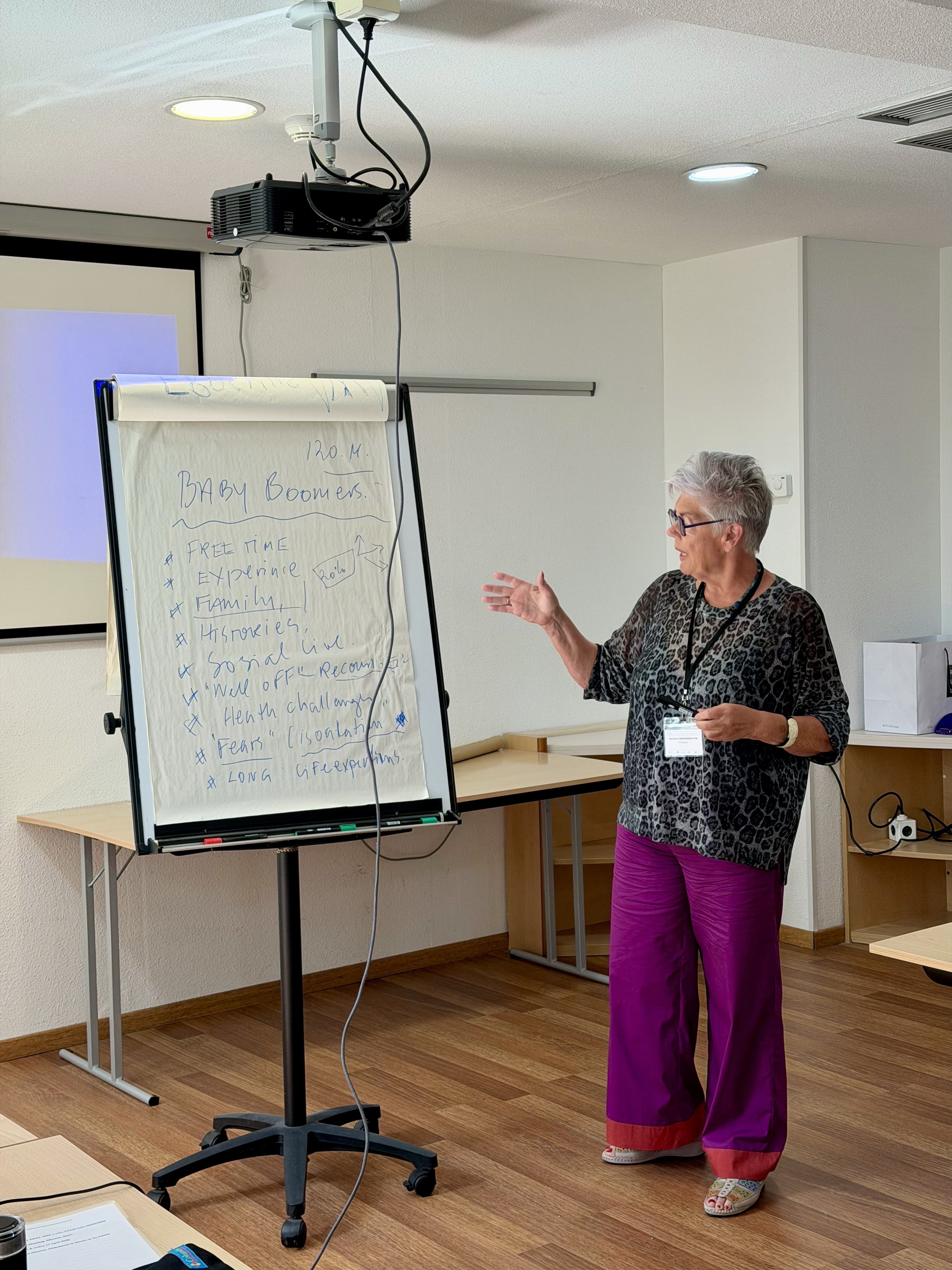 A woman with short gray hair, glasses, wearing a leopard print top and purple pants is giving a presentation in a room. She is gesturing with her left hand while holding a marker in her right hand. There is a whiteboard with handwritten notes, a proj