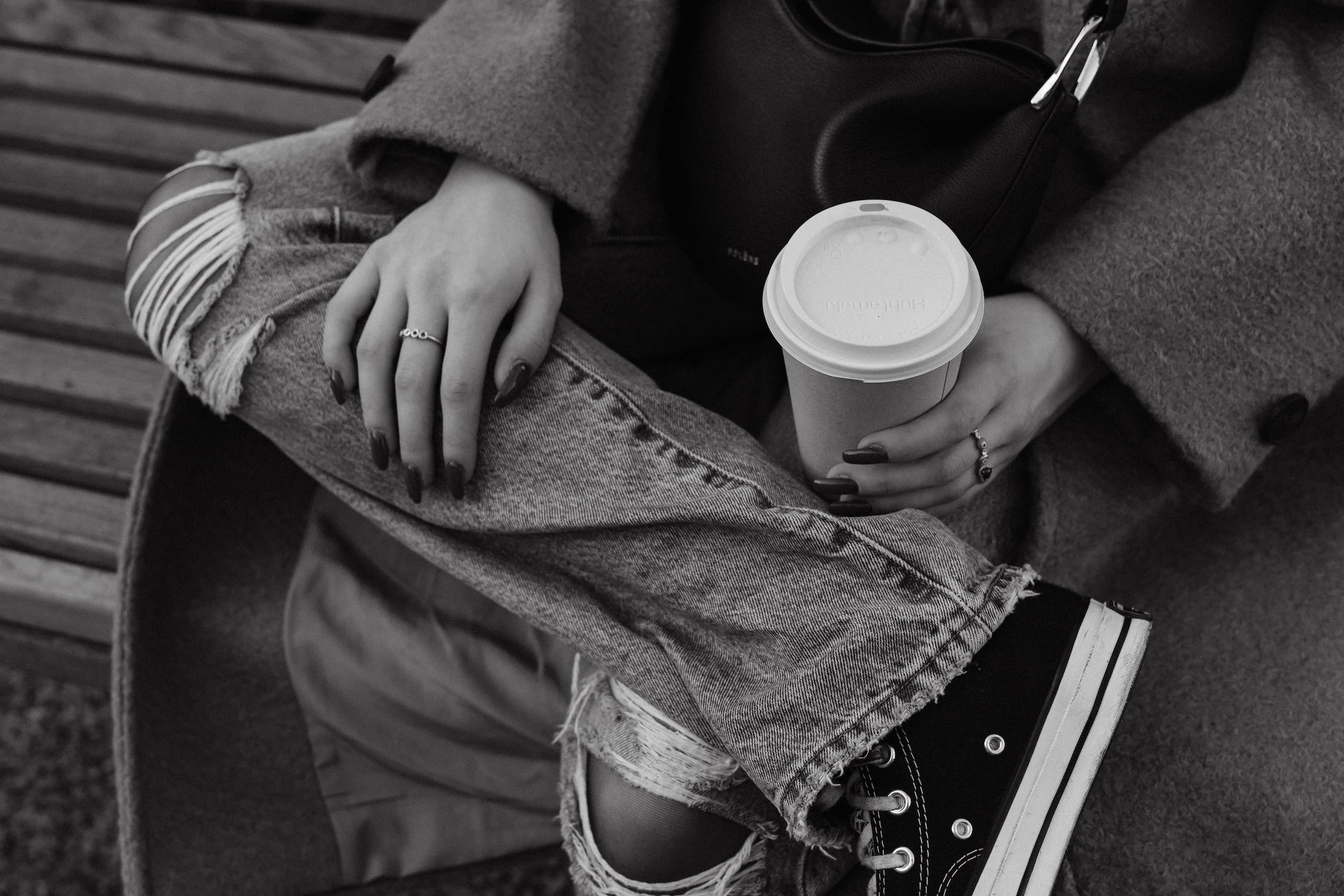 A person sitting outdoors on a bench, wearing ripped jeans, a coat, and a ring, holding a to-go coffee cup and a smartphone.