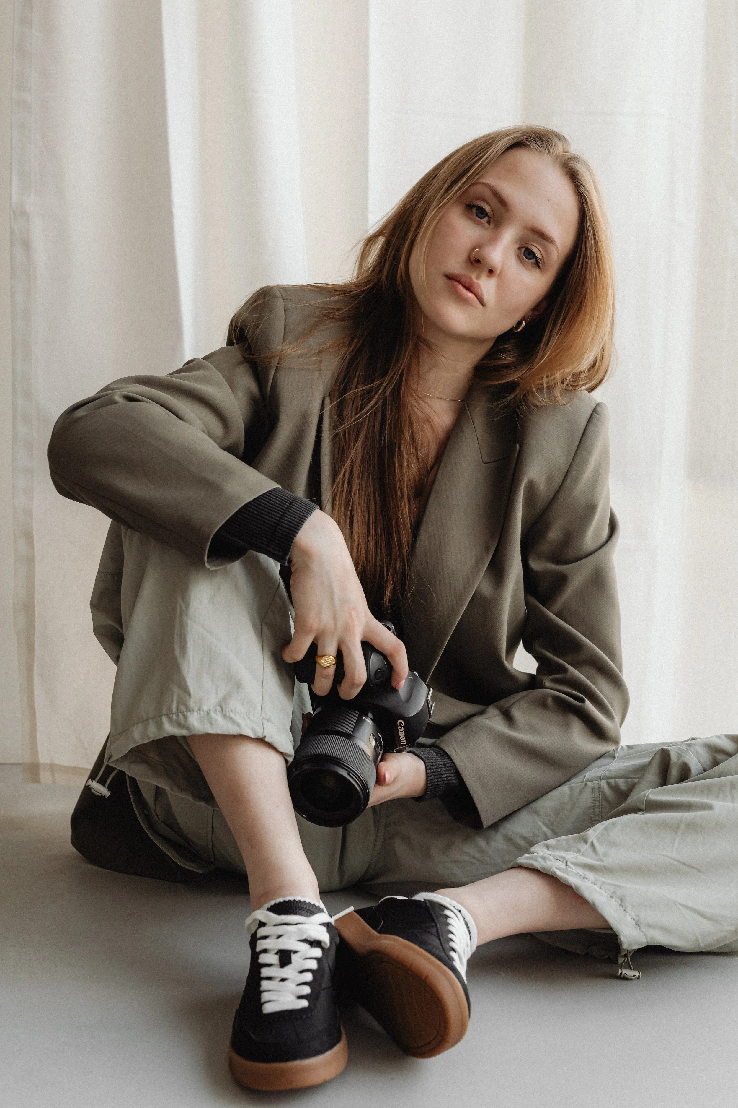 A young woman with long reddish-brown hair and pale skin sitting on the floor with a camera in her hand, wearing a greenish-brown blazer, tan shorts, and black sneakers, in front of a white curtain.