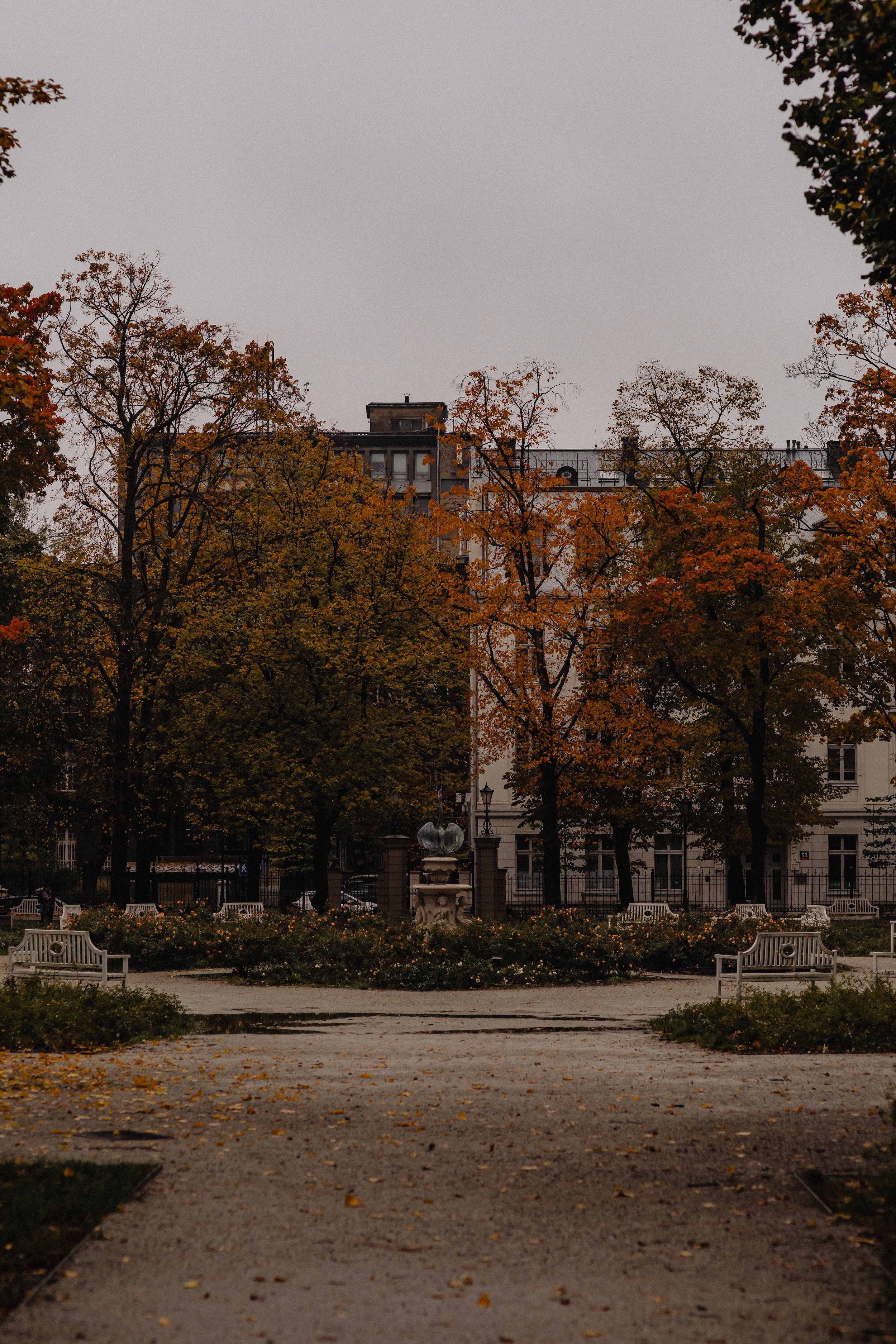 A park with orange and yellow autumn trees, benches, and a stone sculpture in the center. Buildings are visible behind the trees, and the sky is cloudy.