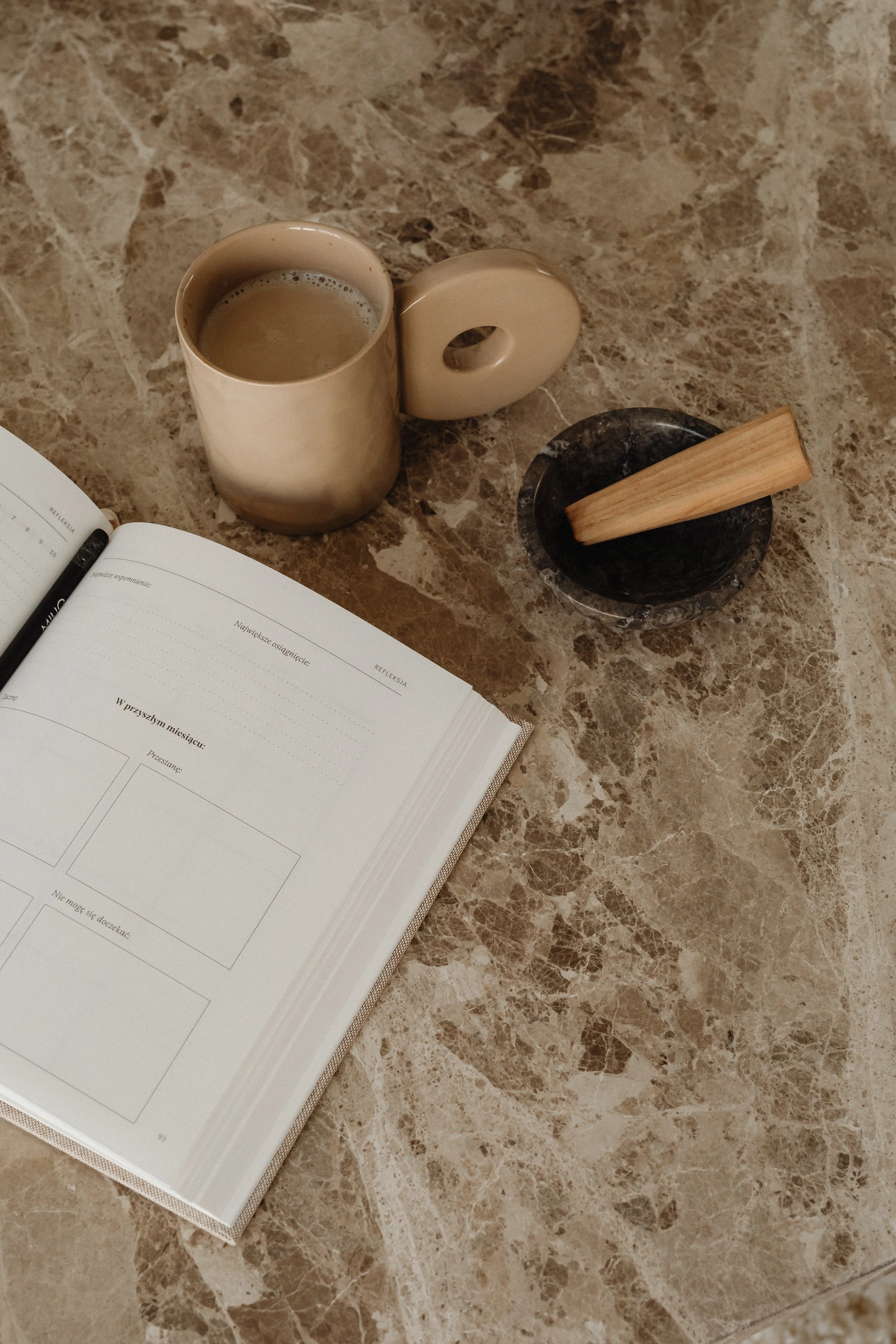 Open notebook and pen on a marble table with a beige mug filled with a hot beverage, a black dish, and a wooden pestle.