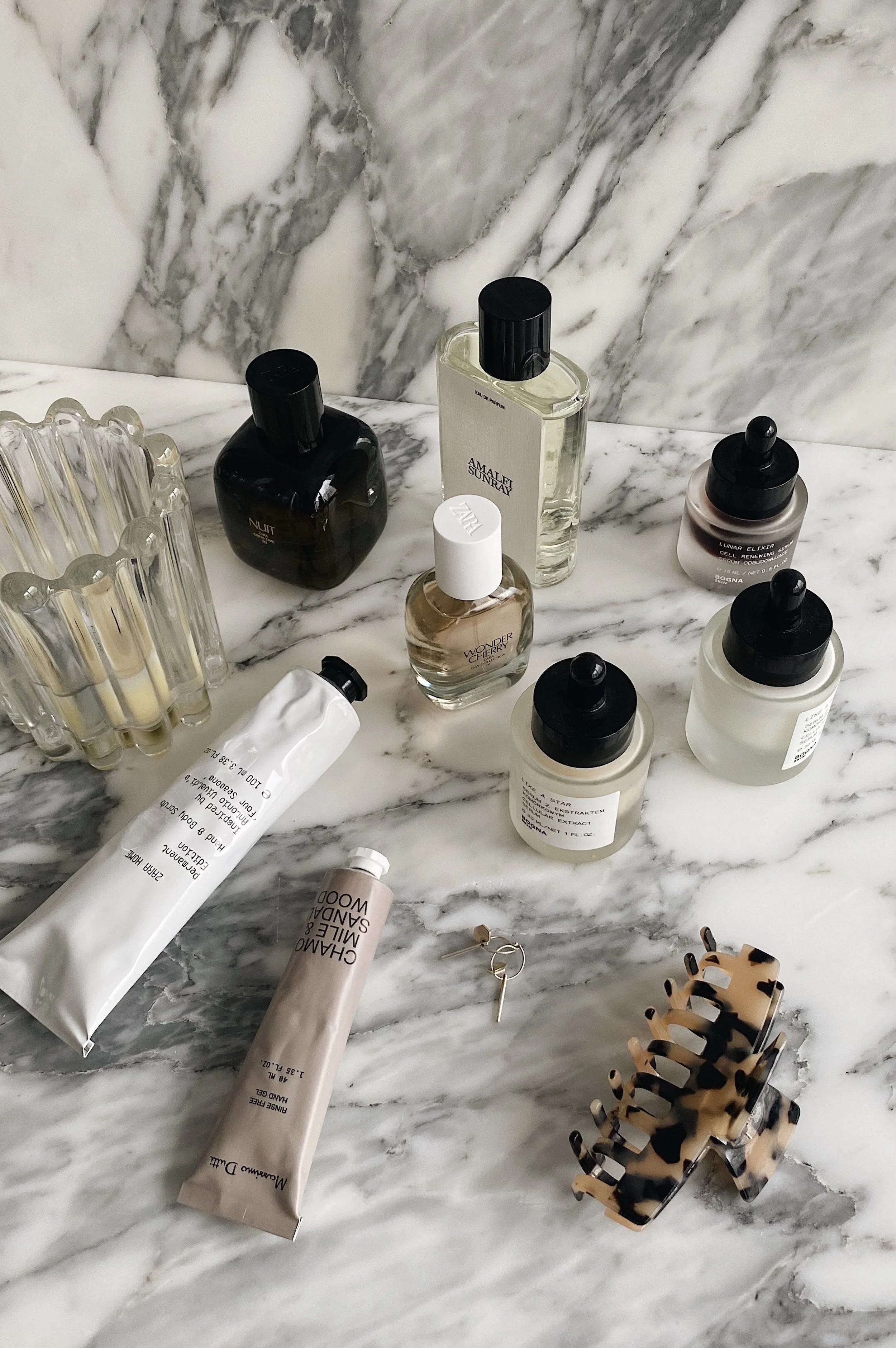 A marble countertop with various skincare and cosmetic bottles, cream tubes, a hair claw clip, a hairpin, and a tortoise shell hair clip.