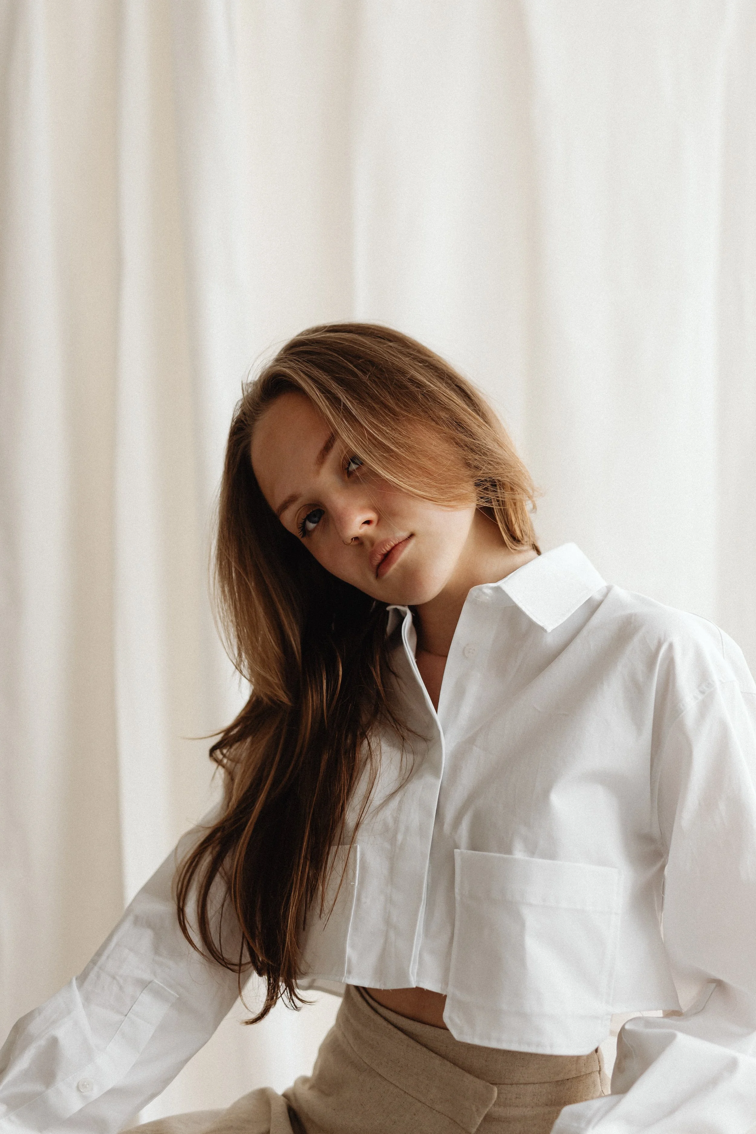 A young woman with long wavy brown hair and light skin, wearing a white cropped shirt and beige high-waisted pants, standing against a plain curtain background.