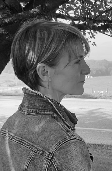 A woman with short hair and earrings stands outdoors near a tree, looking to the right, with a road and landscape in the background.