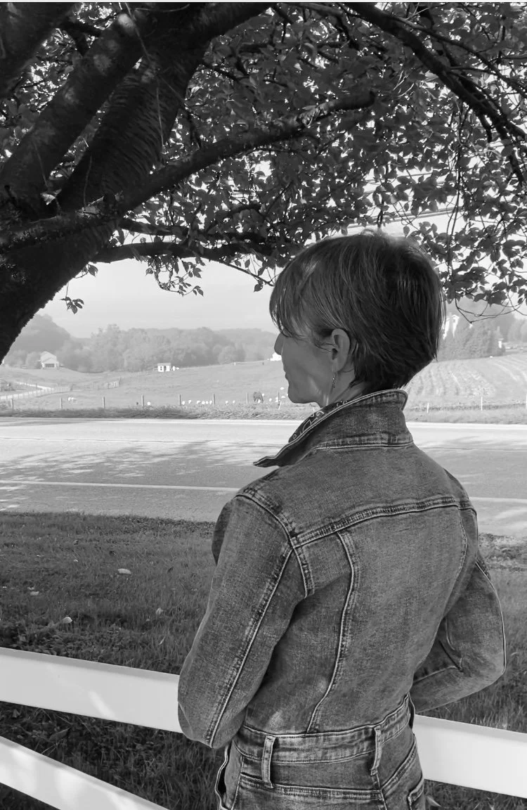 A woman with short hair wearing a denim jacket stands outdoors under a tree, looking into the distance at a rural landscape with fields and a distant house.