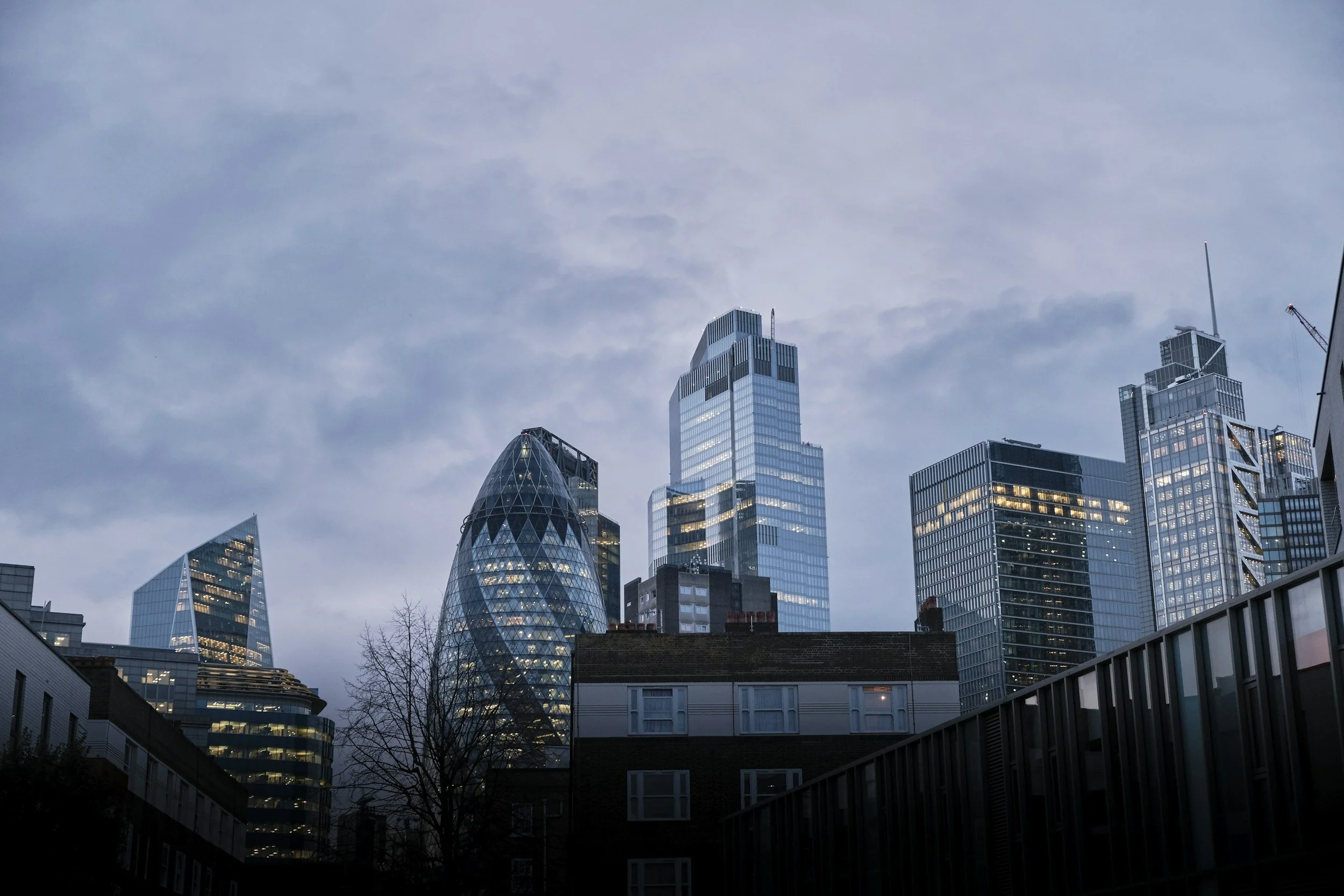 Skyline of modern skyscrapers in a city with cloudy sky in the background, some buildings with reflective glass windows, and construction cranes visible.